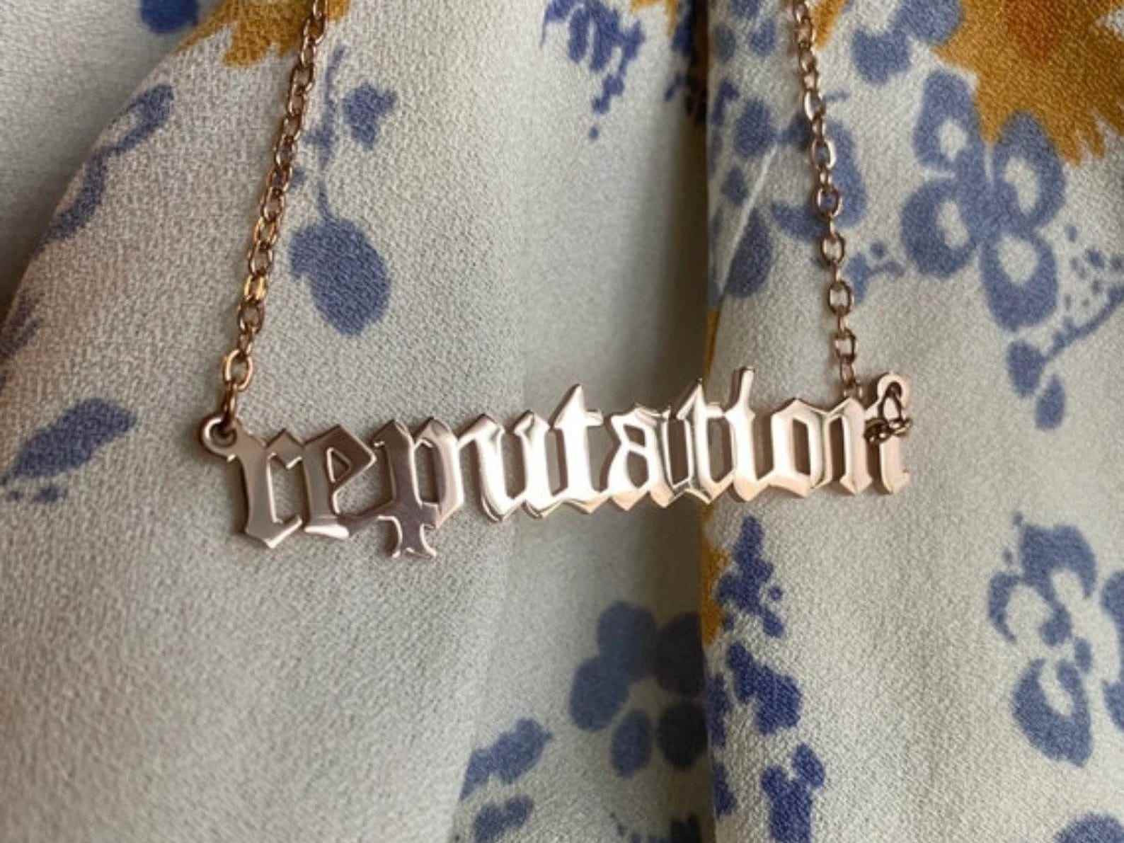 Eine goldene Halskette mit dem Albumtitel „Reputation“ von Taylor Swift auf einer geblümten Decke