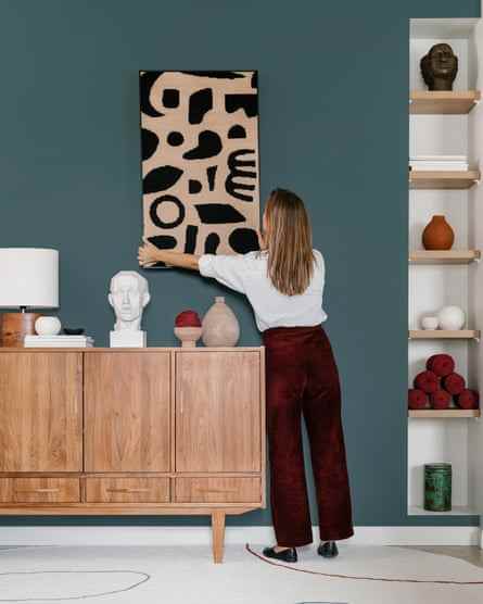 Rückansicht einer nicht erkennbaren Designerin, die ein Bild mit abstraktem Ornament an einer bemalten Wand aufhängt, während sie an der kreativen Innenarchitektur einer modernen Wohnung arbeitet