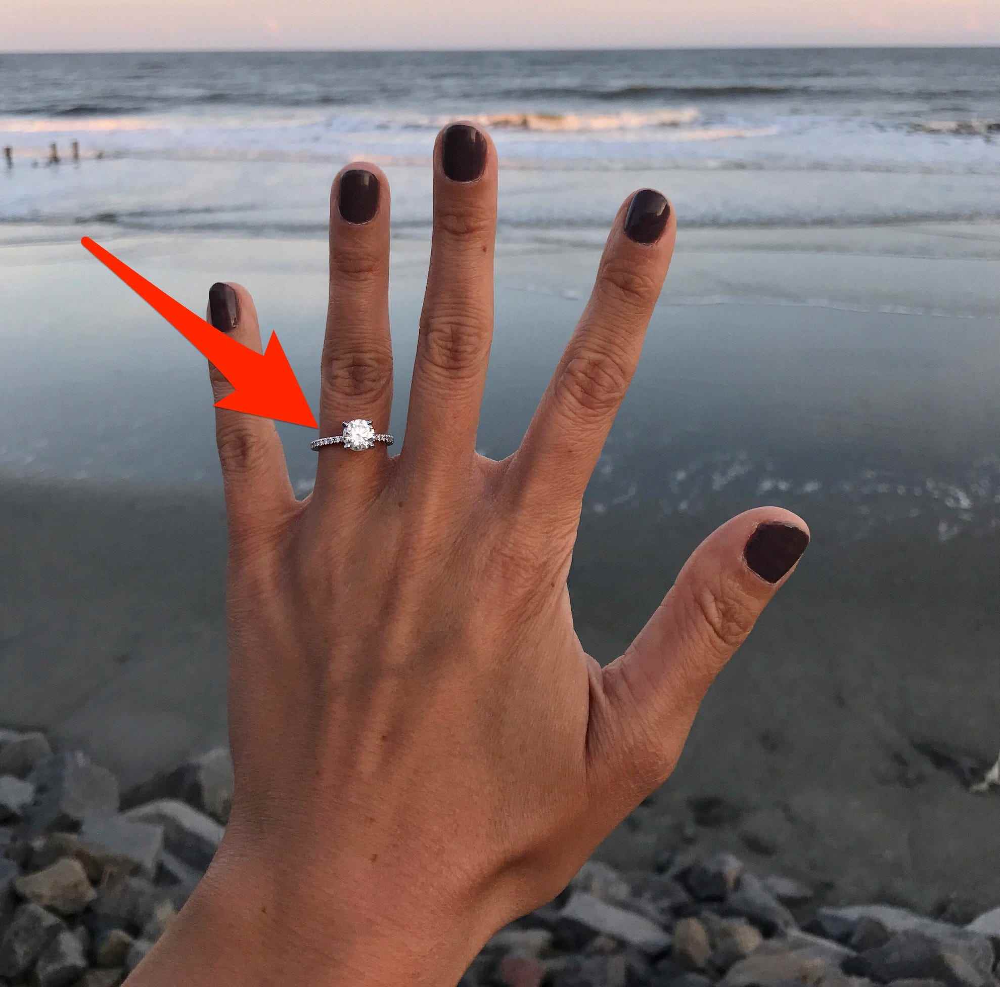 Ein roter Pfeil zeigt auf einen Verlobungsring an der Hand einer Frau, die ihn vor dem Ozean hochhält.
