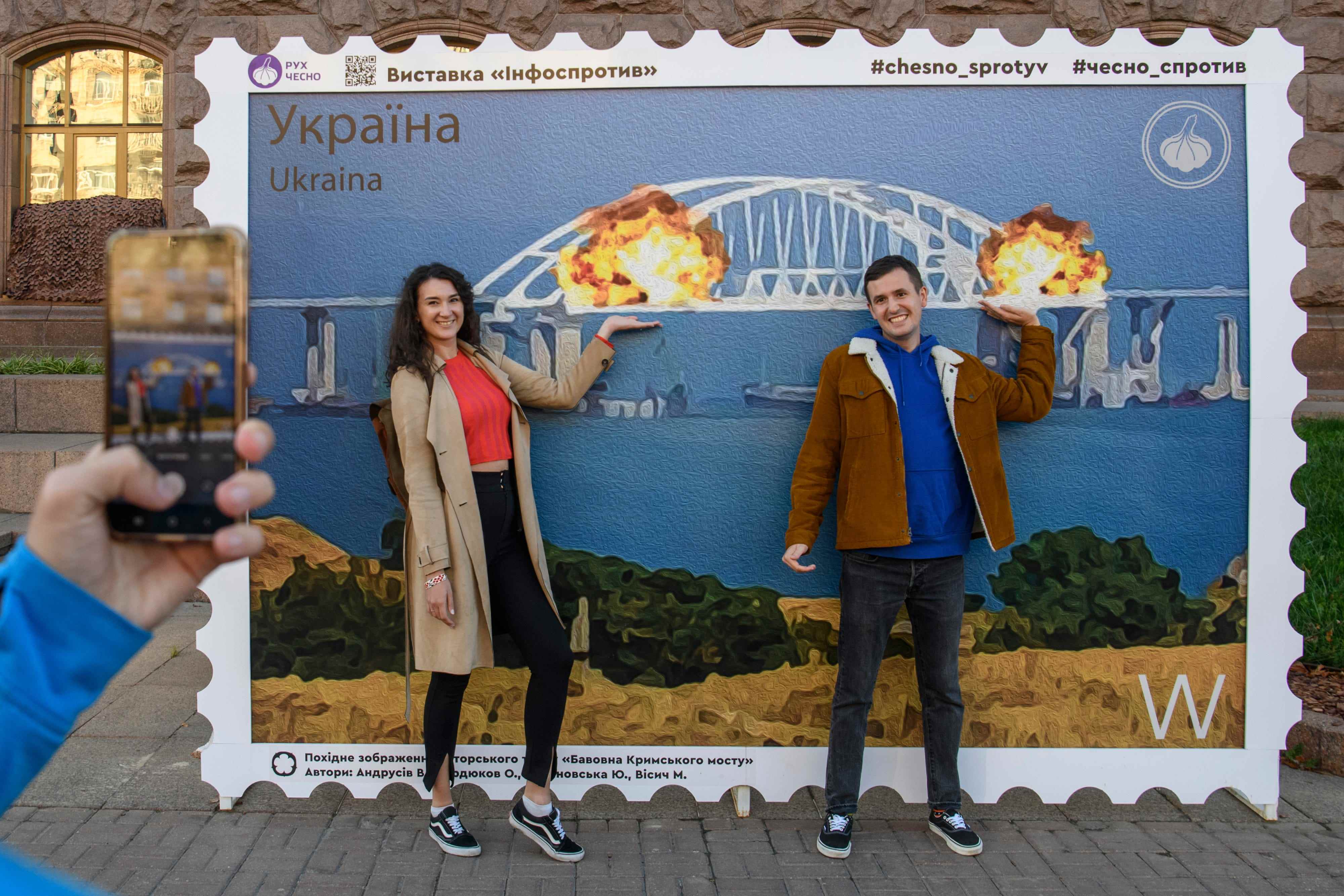 Menschen posieren für Fotos und machen Selfies vor der großen Plakatform einer Briefmarke, die die brennende Krim-Kertsch-Brücke in Kiew, Ukraine, am 8. Oktober 2022 darstellt.