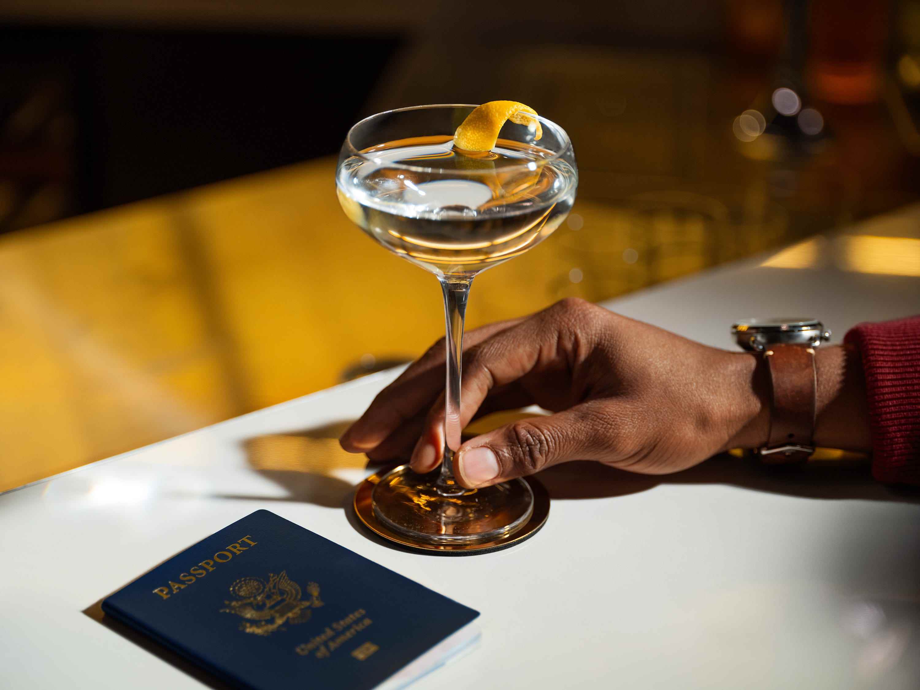Ein Cocktail, der von einer Hand auf einem Tisch neben einem US-Pass in der Chase Sapphire Lounge von The Club in Hongkong gehalten wird.