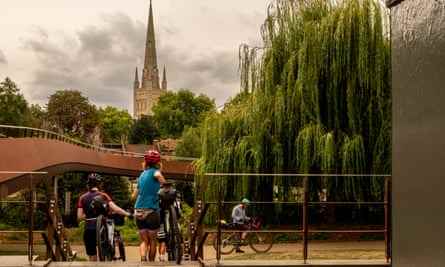 Eine Gruppe von Radfahrern nimmt ihre Fahrräder über eine Brücke über den Fluss nahe dem Zentrum von Norwich.  Ein Domturm ist in der Ferne.