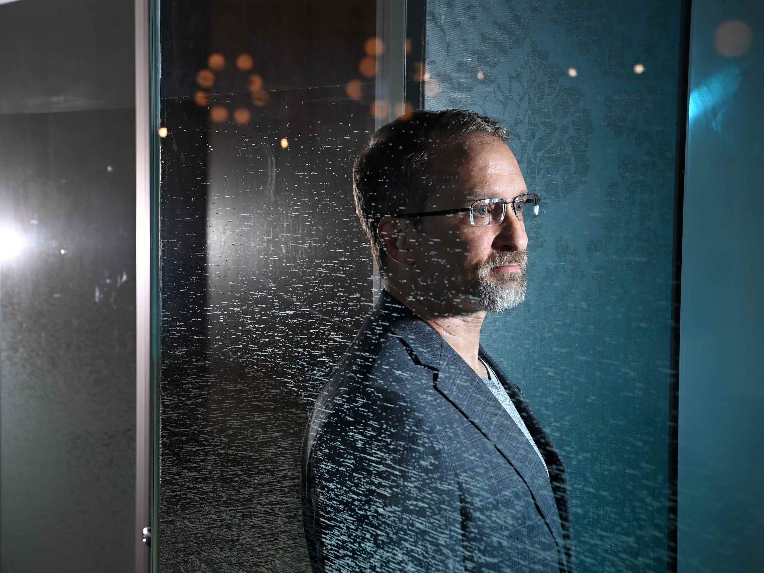 Ein Mann mit grauem Haar, Bart und Brille steht hinter einer Glaswand und starrt in die Ferne.