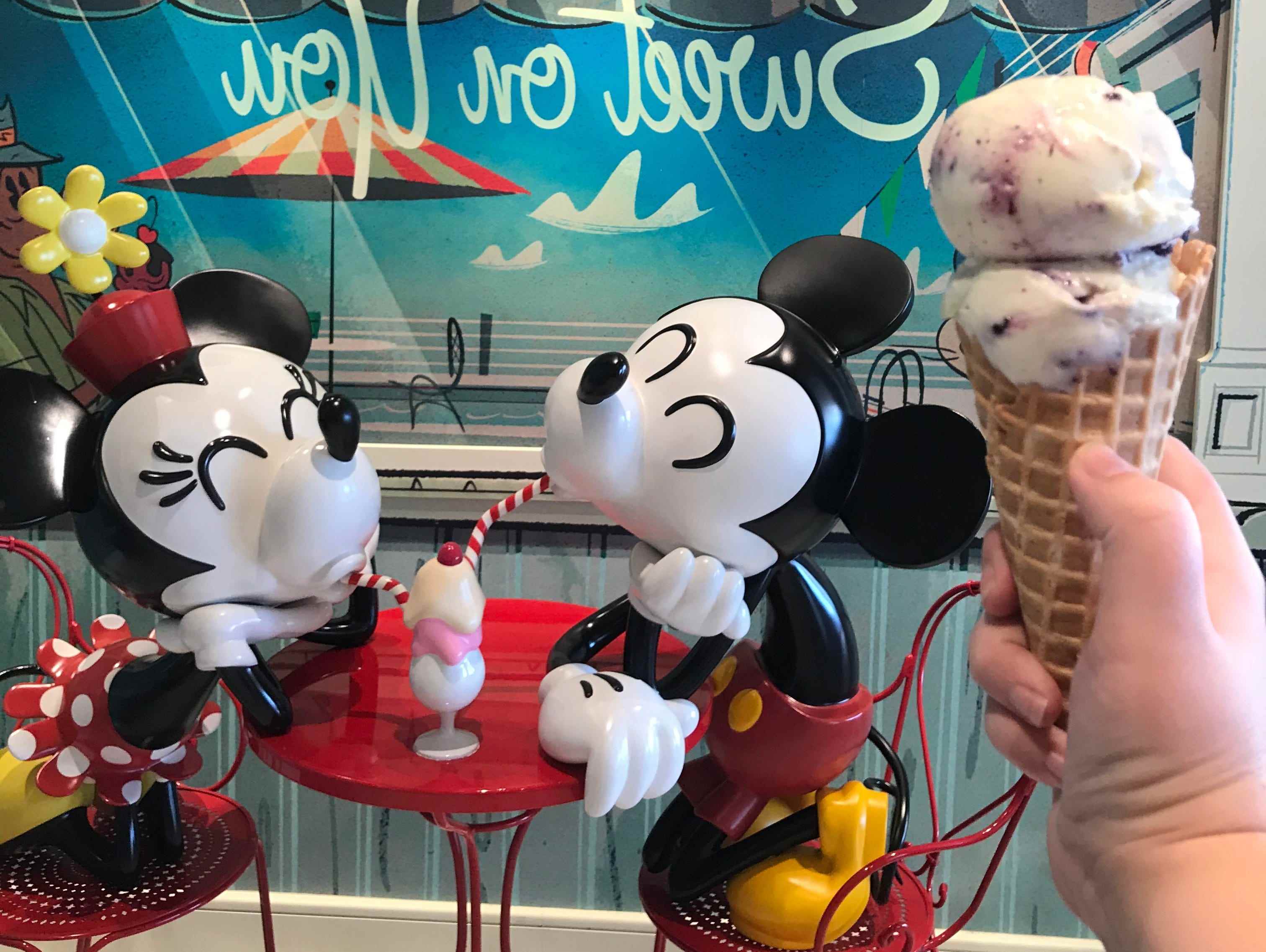 Hand mit Kegel voller Eiskugeln vor einem Eisladen mit Mickey- und Minnie-Statue