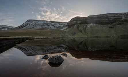 Reflexionen von Bergen in einem See, in dessen Mitte ein großer Felsen sitzt, in Llyn Y Fan Fach, Carmarthenshire, Wales.