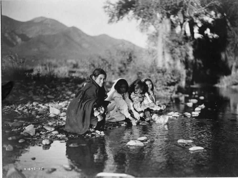 Vier Taos-Kinder hocken auf Felsen am Bachrand, Berge im Hintergrund.