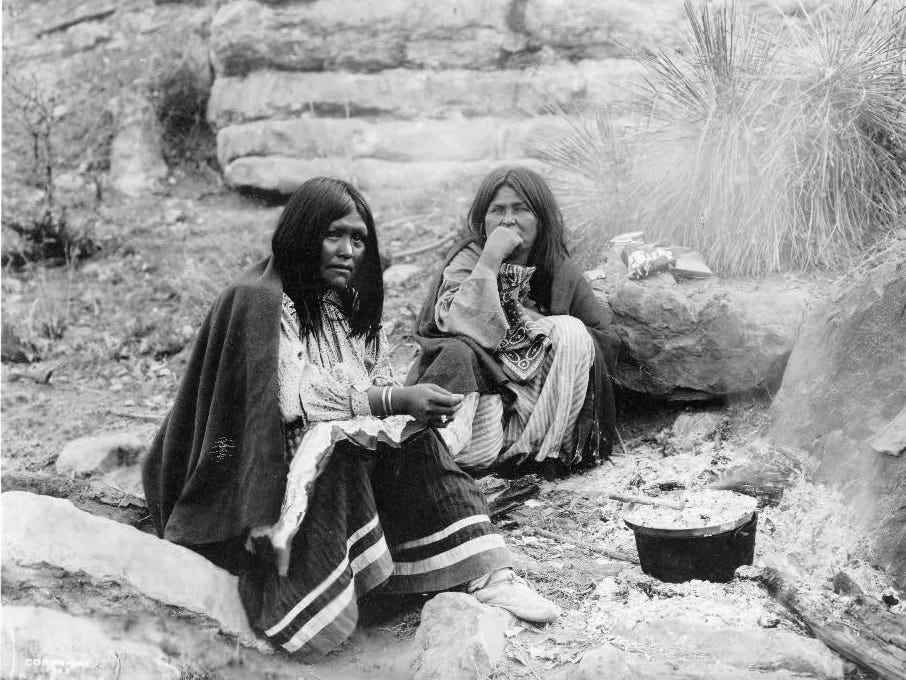 Zwei Apachen-Indianerinnen sitzen vor einem Lagerfeuer, davor ein Kochtopf.