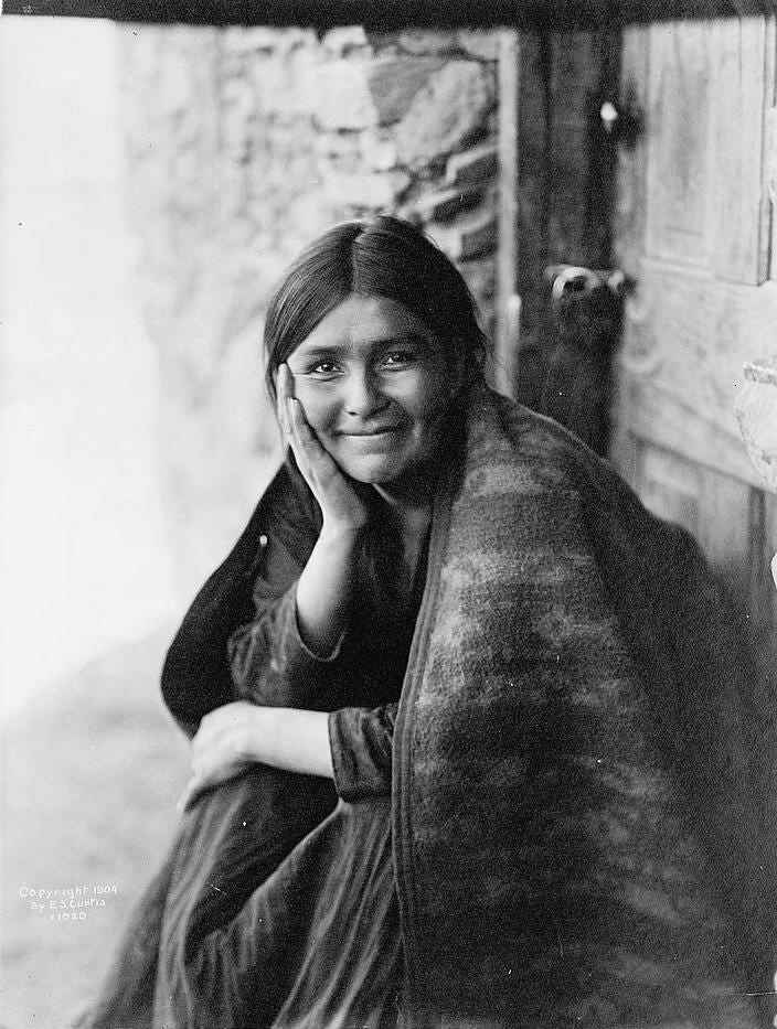 Das Foto zeigt eine Navaho-Frau, die vor der Tür sitzt, die rechte Hand auf der Wange, mit dem Gesicht nach vorne und lächelnd.