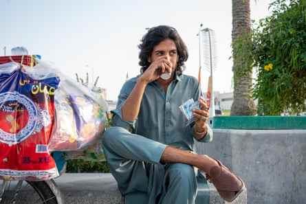 Ein Mann sitzt am Wasser in Doha, Katar, und trinkt Tee aus einem Pappbecher.  Er hat an jedem Finger einen andersfarbigen Ring.