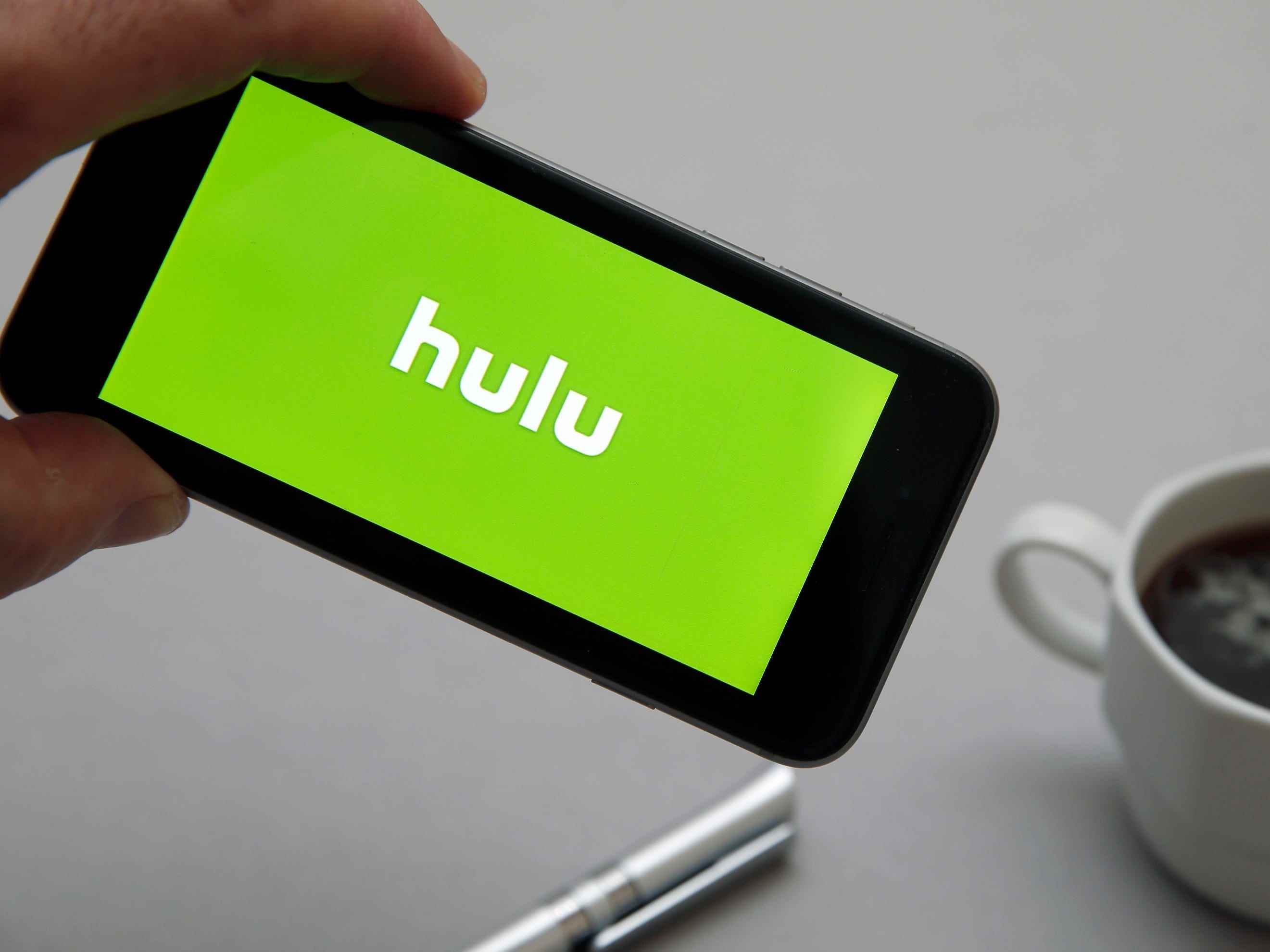 Auf diesem Foto wird das Logo des Hulu-Streamingdienstes auf dem Bildschirm eines Smartphones mit einer Tasse Kaffee auf einem Tisch im Hintergrund angezeigt.
