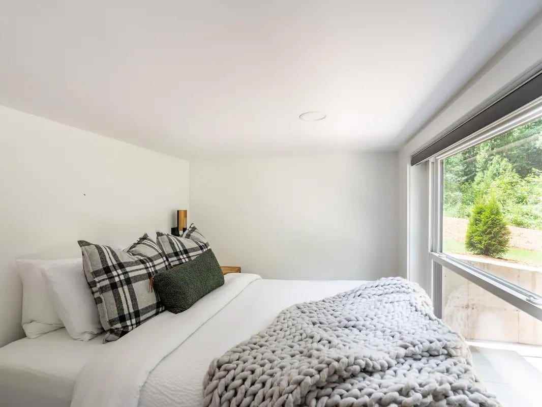 Ein weißes Bett mit grünen Kissen in einem kleinen weißen Zimmer mit Blick auf ein großes Fenster mit Blick auf den Hinterhof