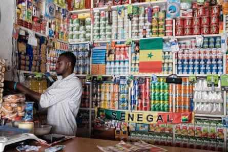 Djibril serviert einem Kunden ein Frühstücksbrötchen, das in seinem Tante-Emma-Laden steht, der mit senegalesischen Flaggen und Schals über den Regalen geschmückt ist.