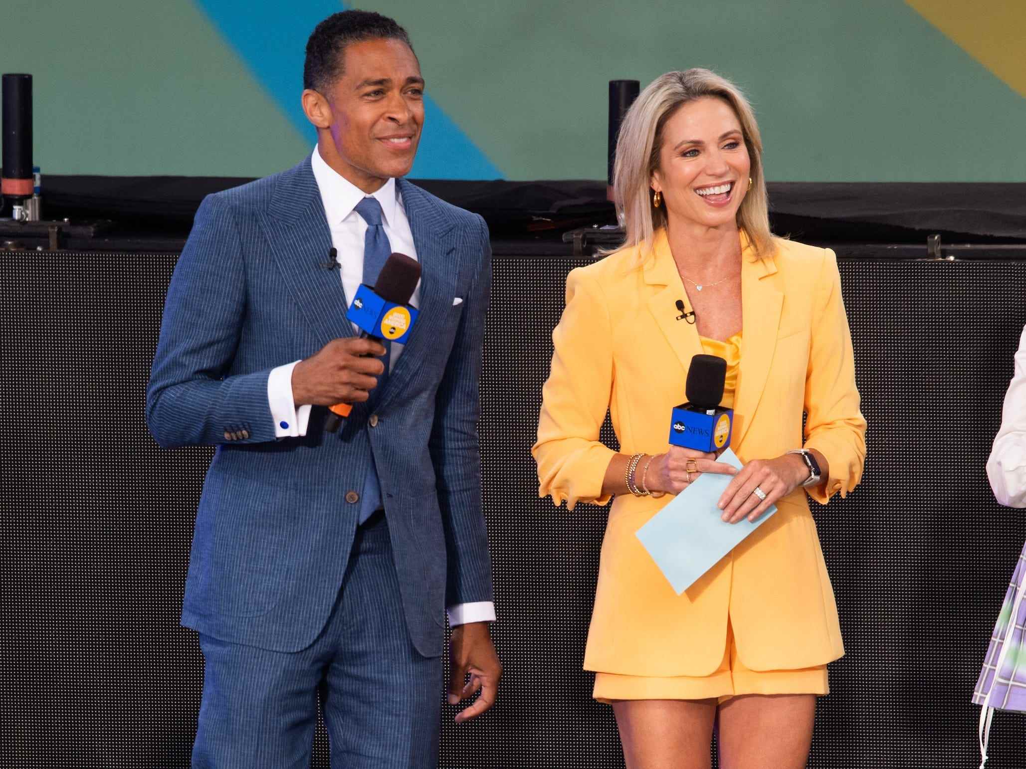 TJ Holmes, in einem blauen Anzug, lächelt, während er neben der „GMA“-Moderatorin Amy Robach, in einem gelben Blazer und Rock, auf der Bühne steht.