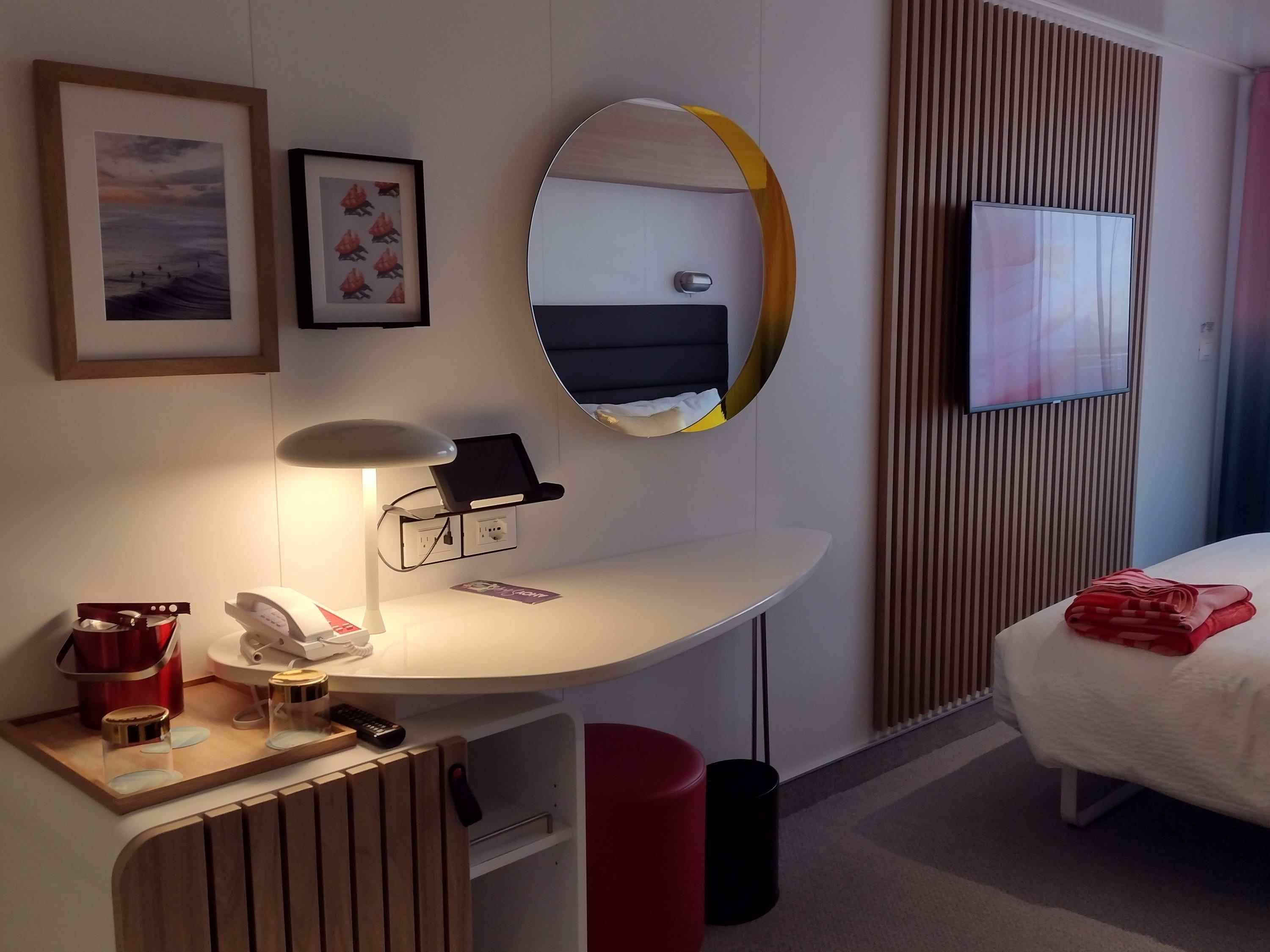 Der Schreibtisch in einer Kreuzfahrtkabine mit Spiegel, Kunstwerken, einem Bett und einem Fernseher.
