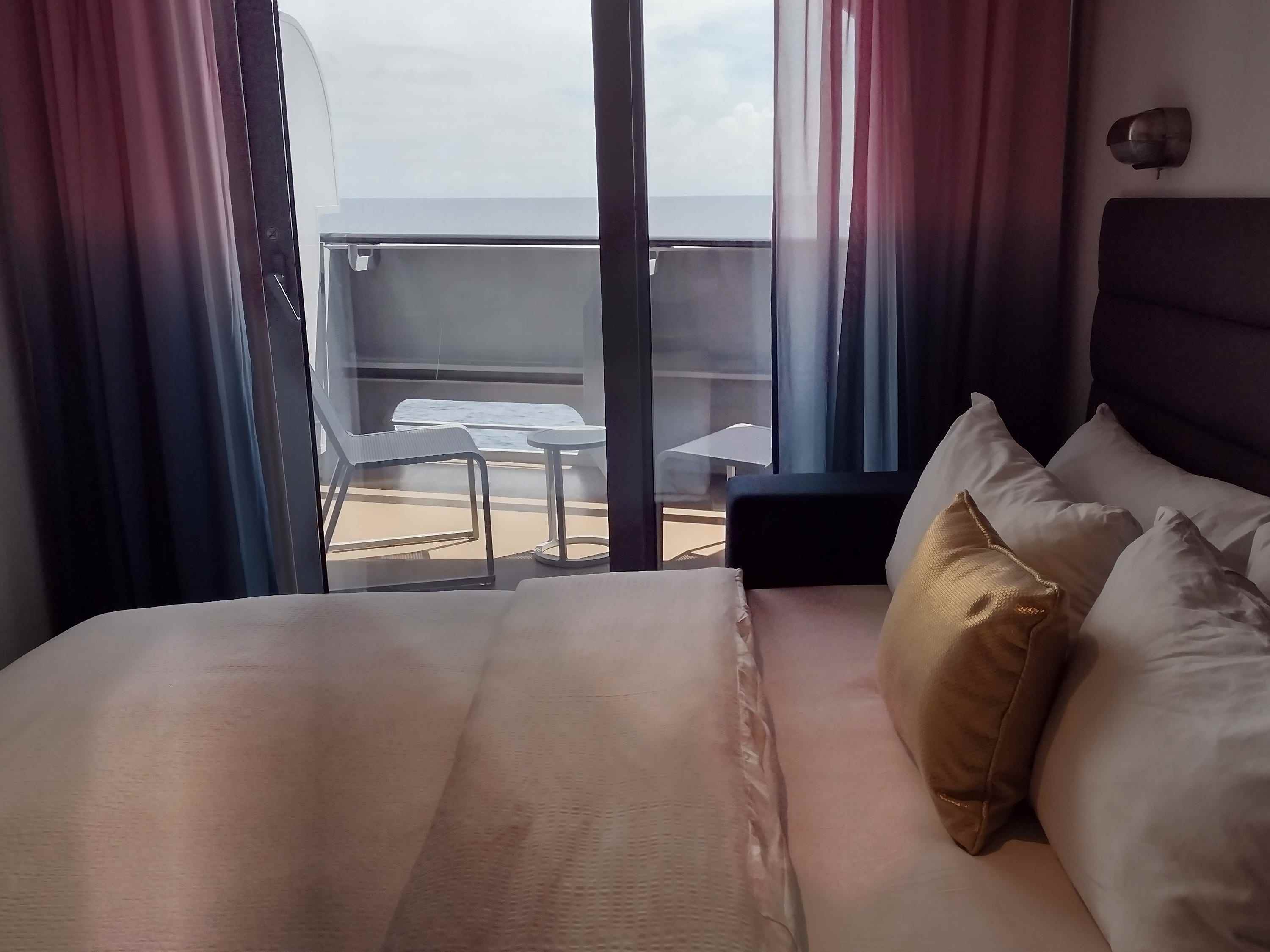 Ein Bett auf einem Kreuzfahrtschiff mit offenen Vorhängen, die den Balkon zeigen.