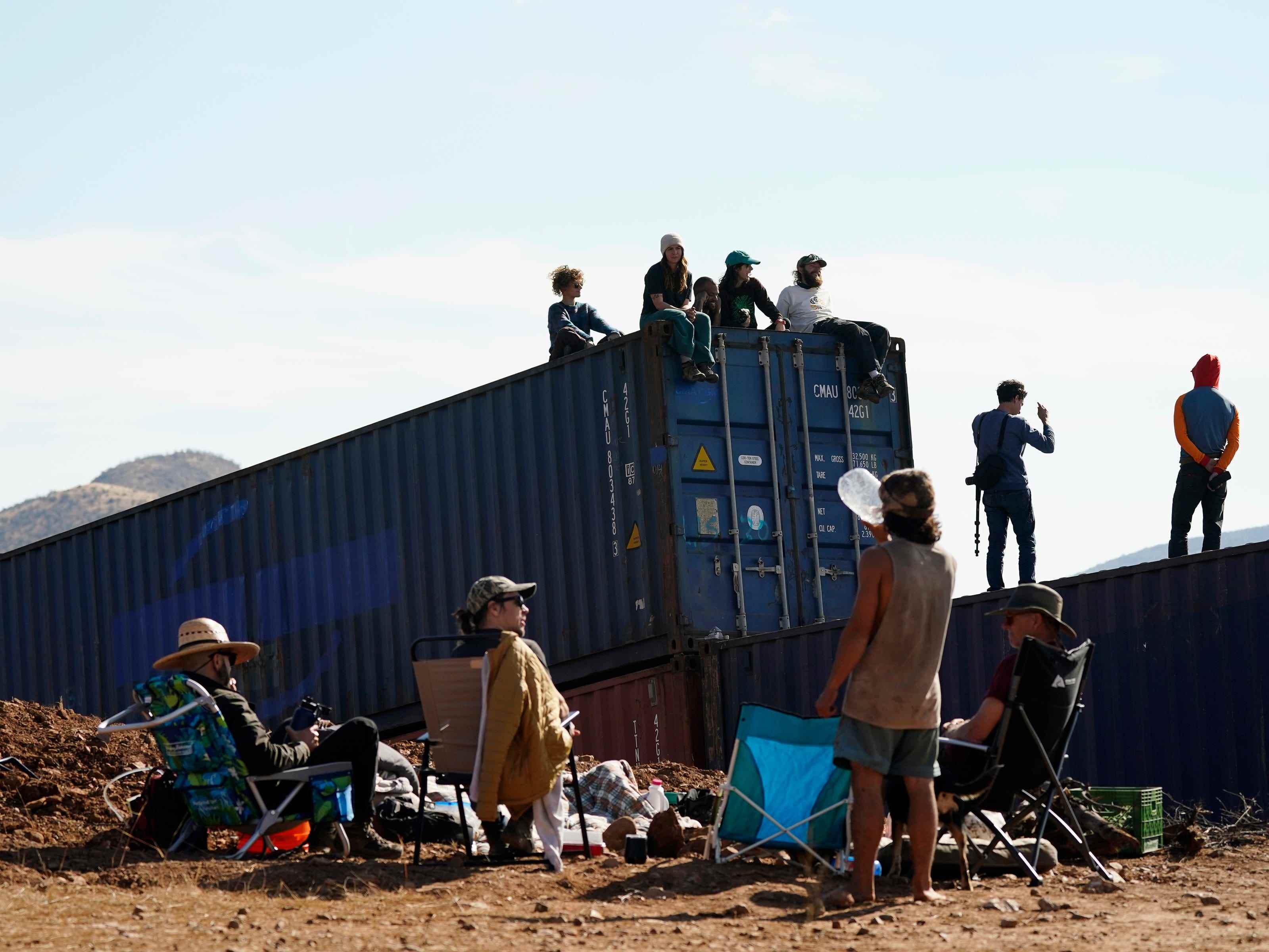 Aktivisten versammeln sich in der Nähe und sitzen auf neu installierten Schiffscontainern entlang der Grenze, die am Donnerstag, den 8. Dezember 2022, im San Rafael Valley, Arizona, eine Mauer zwischen den Vereinigten Staaten und Mexiko errichten.