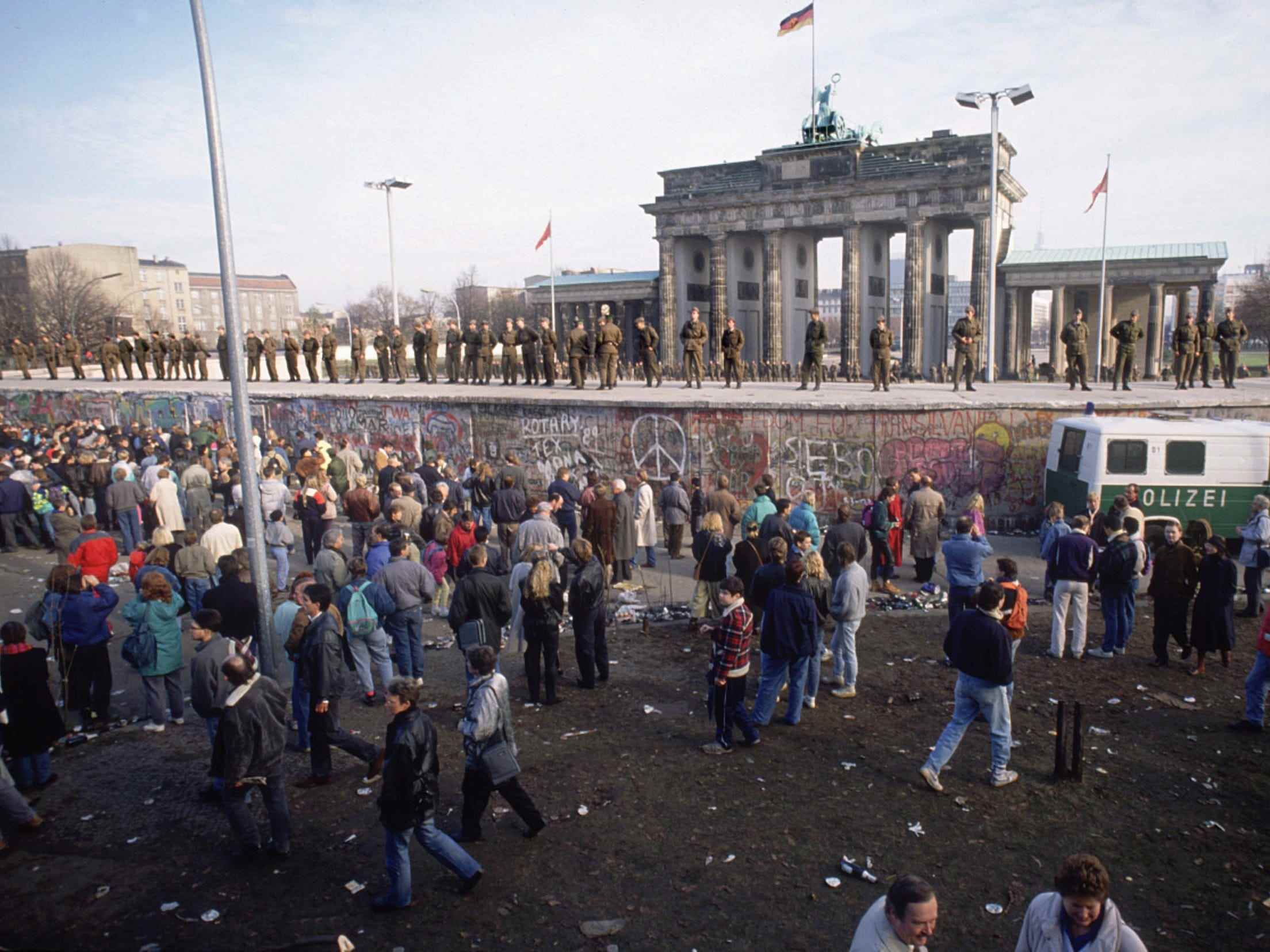 Menschen versammeln sich auf einer Seite der Berliner Mauer, während deutsche Soldaten auf einer erhöhten Plattform vor dem Brandenburger Tor stehen