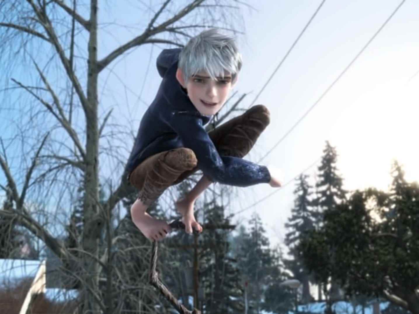 Jack Frost, ein junger Mann mit silbernem Haar, nackten Füßen und einem blauen Hoodie, der wie ein Teenager aussieht.  er steht oben auf einem Stab, nachdem er gerade einen Schneeball geworfen hat