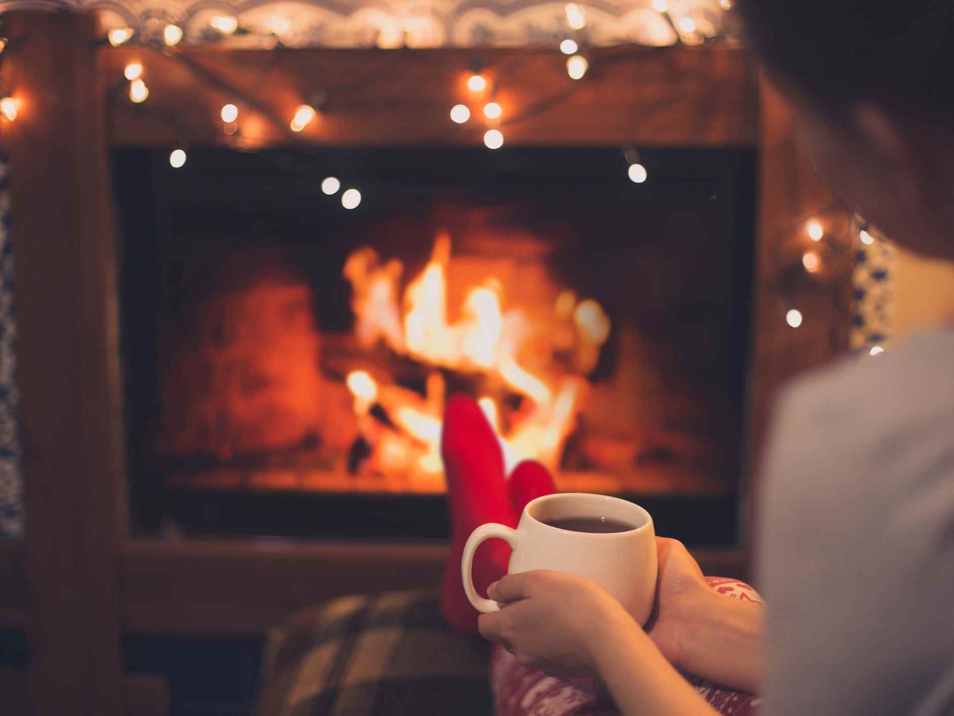 Schließen Sie eine Tasse heißen Tee in den Händen einer Frau, die in der Nähe eines Kamins mit festlichen Weihnachtslichtern in einem gemütlichen Zimmer sitzt.