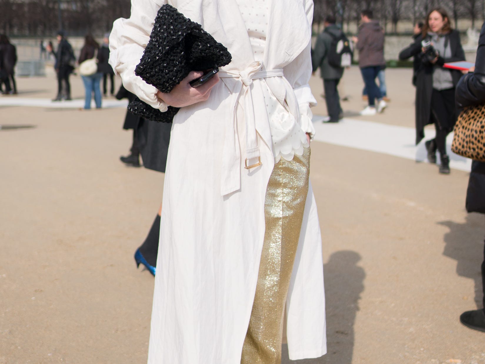 Person, die einen weißen Trenchcoat über einer goldglänzenden Hose und schwarzen Absätzen trägt