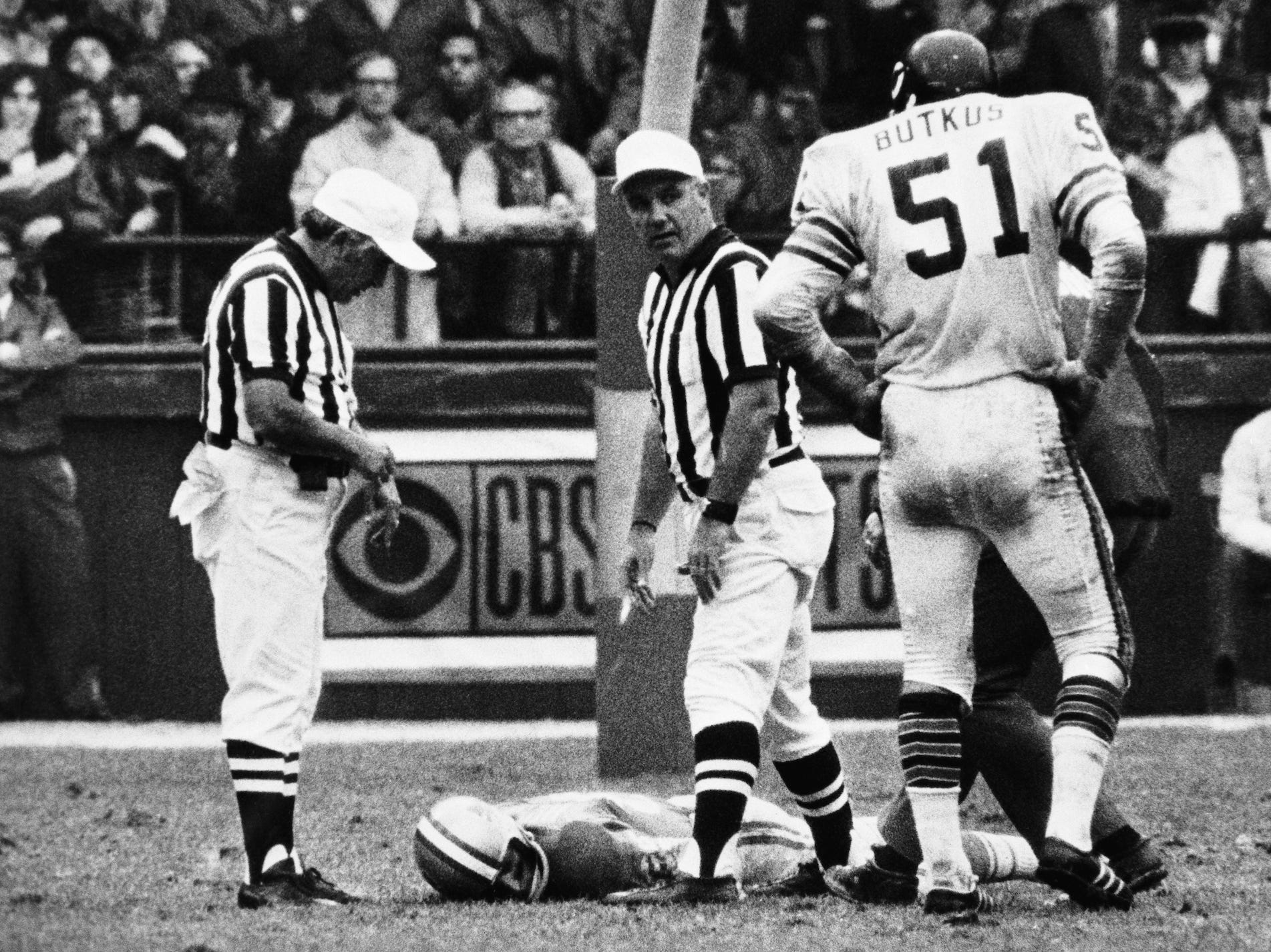 Chuck Hughes liegt mit dem Gesicht nach unten auf dem Feld, während zwei Schiedsrichter auf einem Schwarz-Weiß-Bild zuschauen.