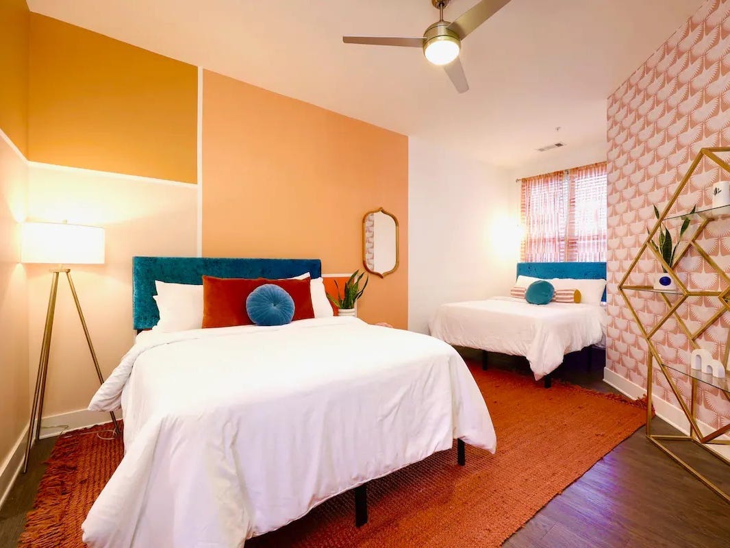 Ein Schlafzimmer mit orangefarbenen und gelben Wänden.  Und ein Bett mit weißer Bettwäsche und blauen und orangefarbenen Kissen.