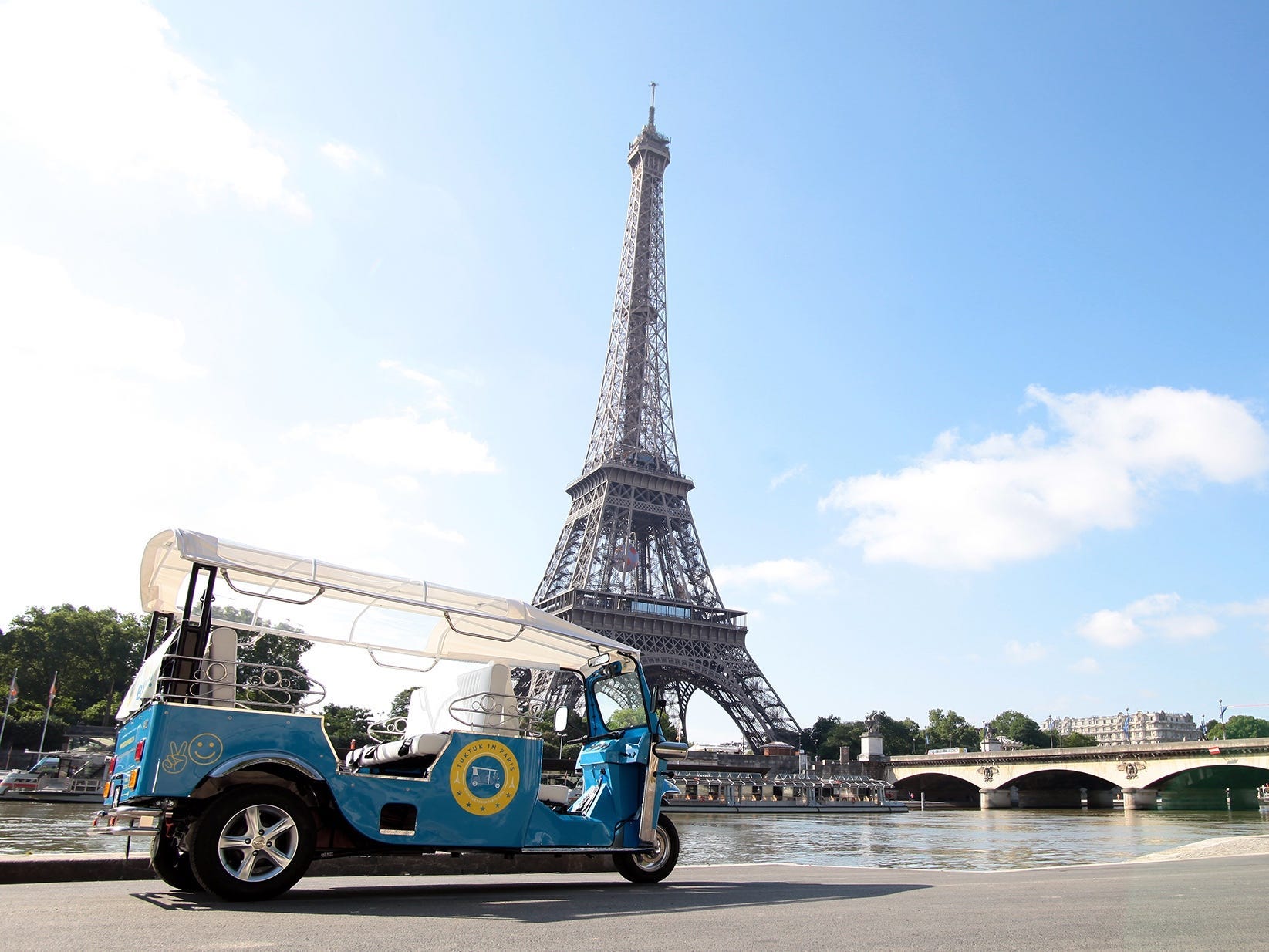 Vor dem Eiffelturm parkt ein kleines, blaues Gefährt, ähnlich einem Golfcart