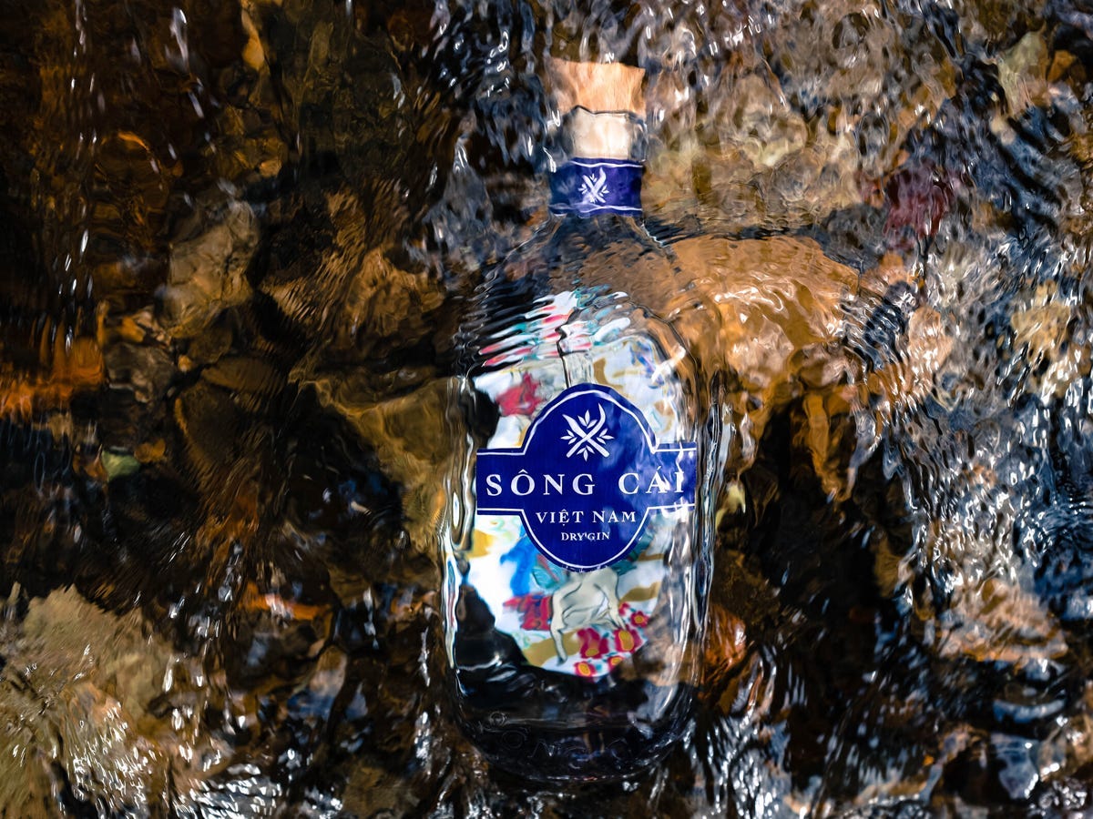 Eine durchsichtige Flasche mit dem Text „song cai vietnam dry gin“ auf einem blauen Etikett, die Flasche liegt in einem Wasserstrahl