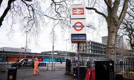 Ein Mann in einem Warnschutz-Arbeitsanzug, der neben einem Schild mit dem alten British Rail-Symbol und einem U-Bahn-Symbol auf dem Bürgersteig einer viel befahrenen Straße spaziert