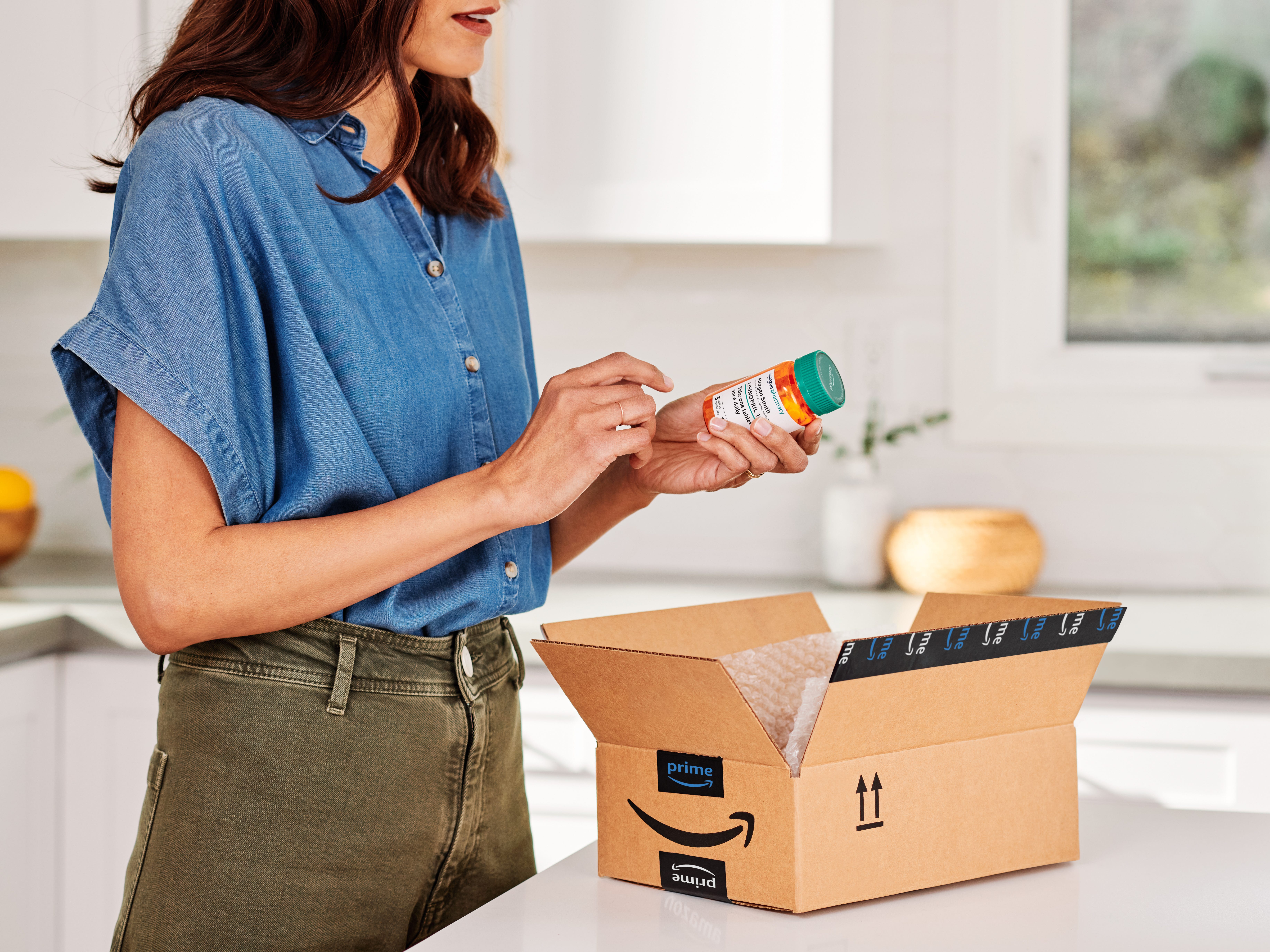 Eine Frau steht mit einer verschreibungspflichtigen Flasche in der Hand über einer Kiste von Amazon RxPass durch Amazon Pharmacy.
