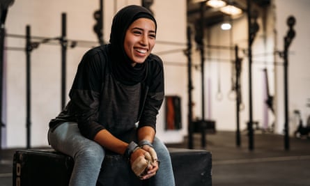 Fröhliche junge muslimische Frau in Hijab und Sportbekleidung, die auf einer Fitbox sitzt, während sie während des Trainings im modernen Fitnessstudio mit Sportgeräten Pause macht