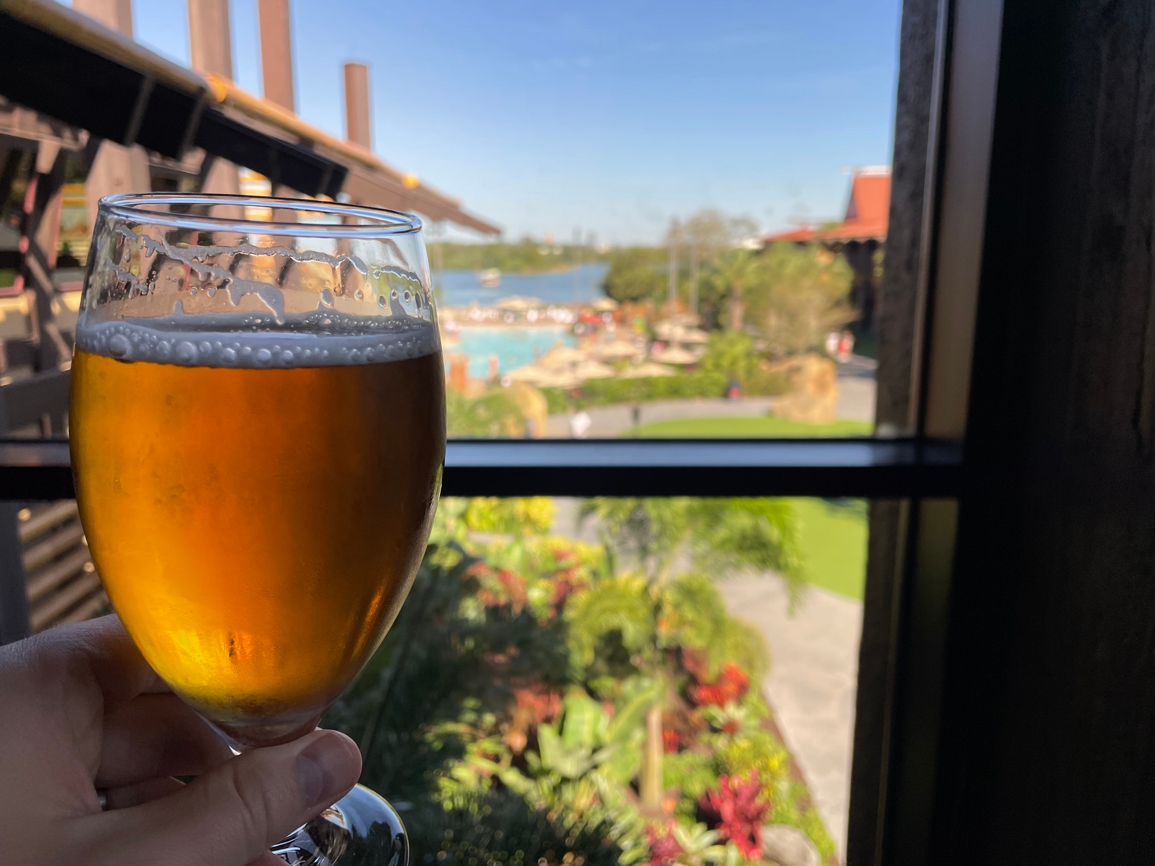 Hand mit einem Glas Bier vor einem Fenster mit Blick auf Disneys polynesisches Resort