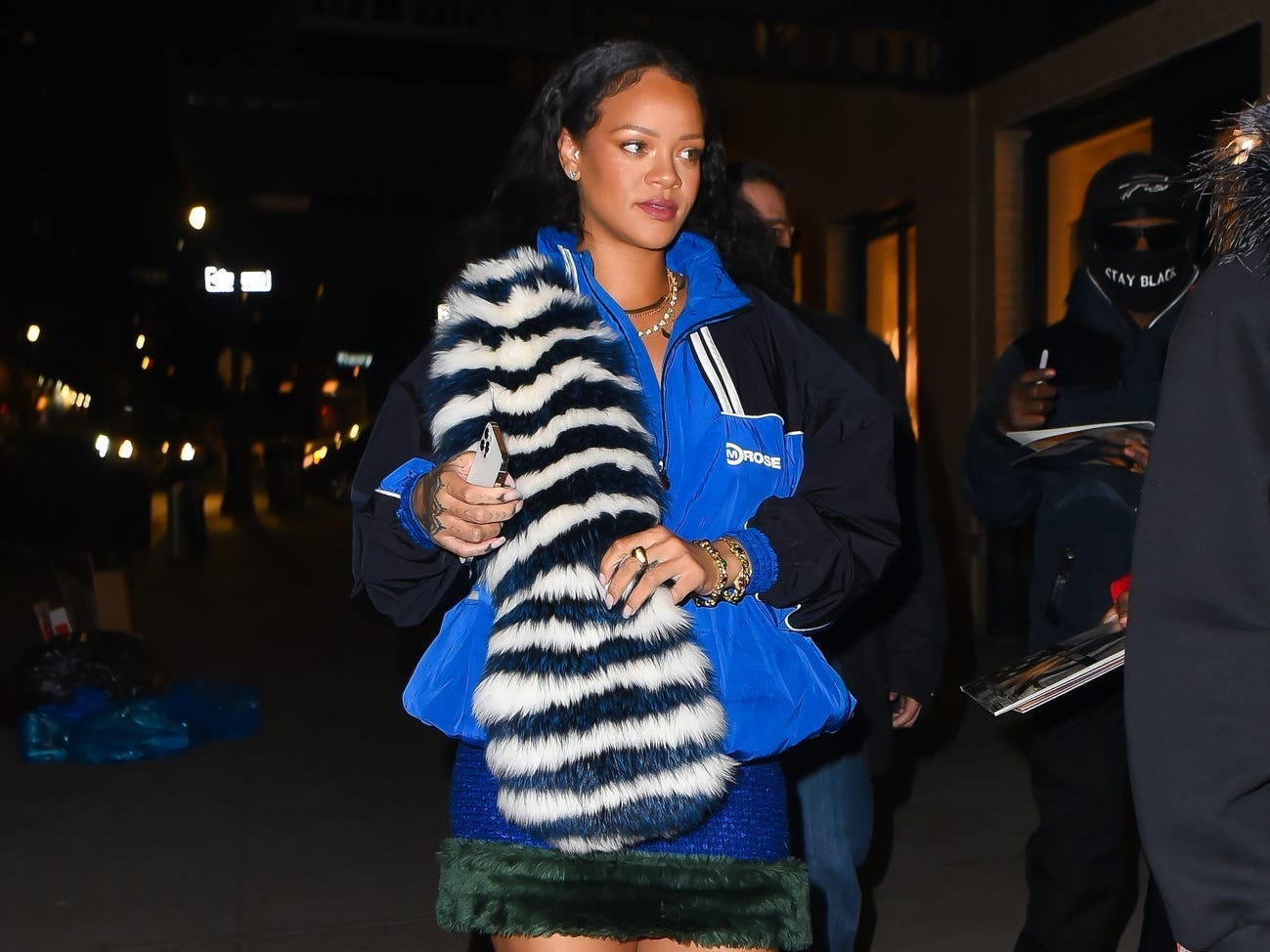 Rihanna in einer blau-schwarzen Windjacke mit einem schwarz-weiß gestreiften Pelzschal über der Schulter.  Sie trägt einen blauen Rock mit dunkelgrünen Verzierungen und weiße Absätze mit Riemen, die bis zu den Knien um ihre Beine gewickelt sind.