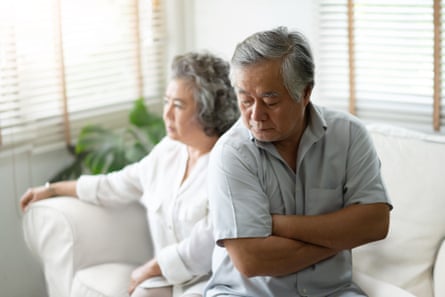 Unglücklicher asiatischer Senior, der mit verschränkten Armen auf der Kante des Sofas sitzt, seine Frau sitzt hinter ihm und schaut in die Ferne.