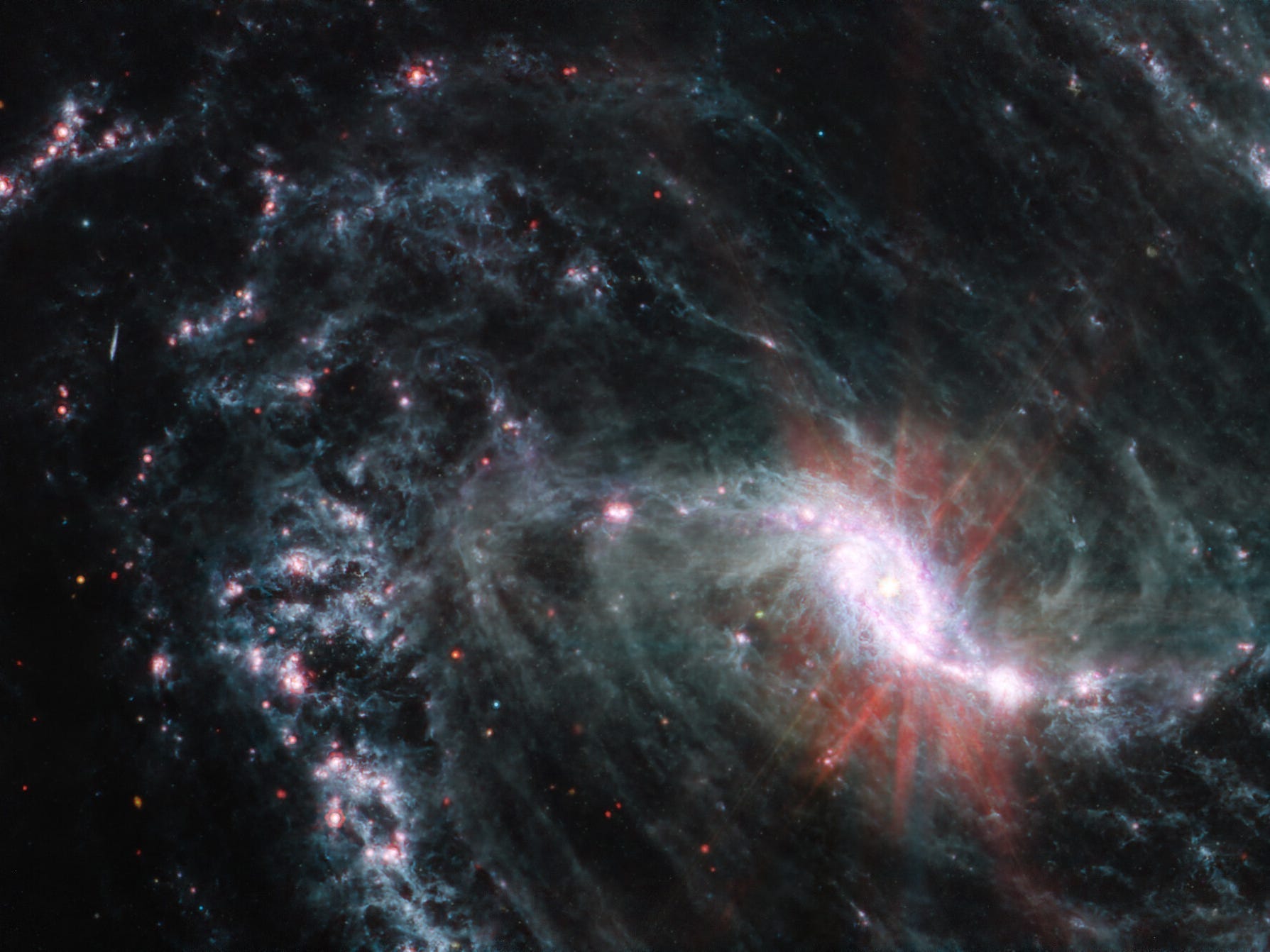 Nahaufnahme einer feinen Spiralgalaxie auf schwarzem Hintergrund.  Es verfügt über einen leuchtenden, hellrosa Kern in einer länglichen ovalen Form.  Rote Streifen scheinen aus dem Kern auszustrahlen.  Der untere rechte Teil der Arme der Galaxie ragt aus dem Rahmen heraus.  Auf dem gesamten Bild stellen vereinzelte kleine rote Punkte Hintergrundgalaxien dar.