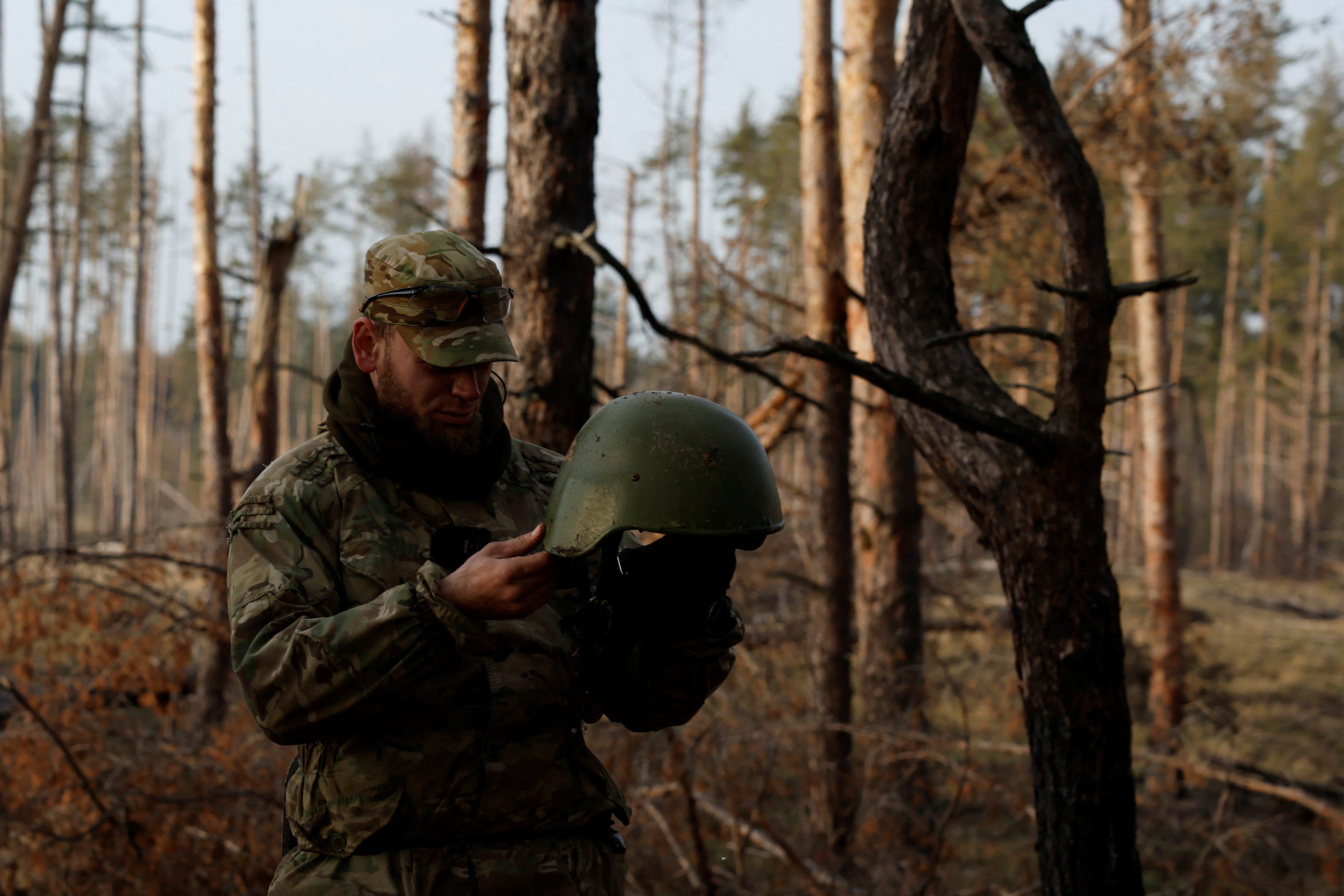 Ein Mann in Camouflage steht im Wald und hält einen Helm in der Hand.