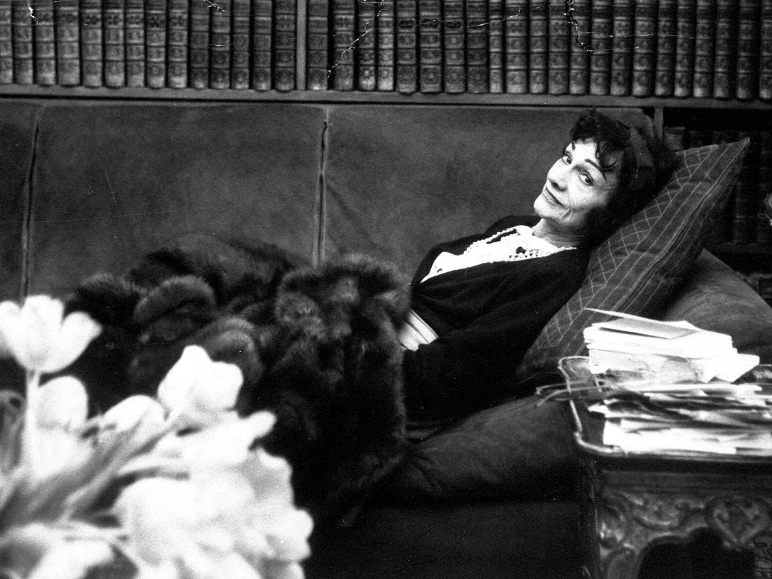 Coco Chanel räkelt sich auf einer Couch vor Bücherregalen