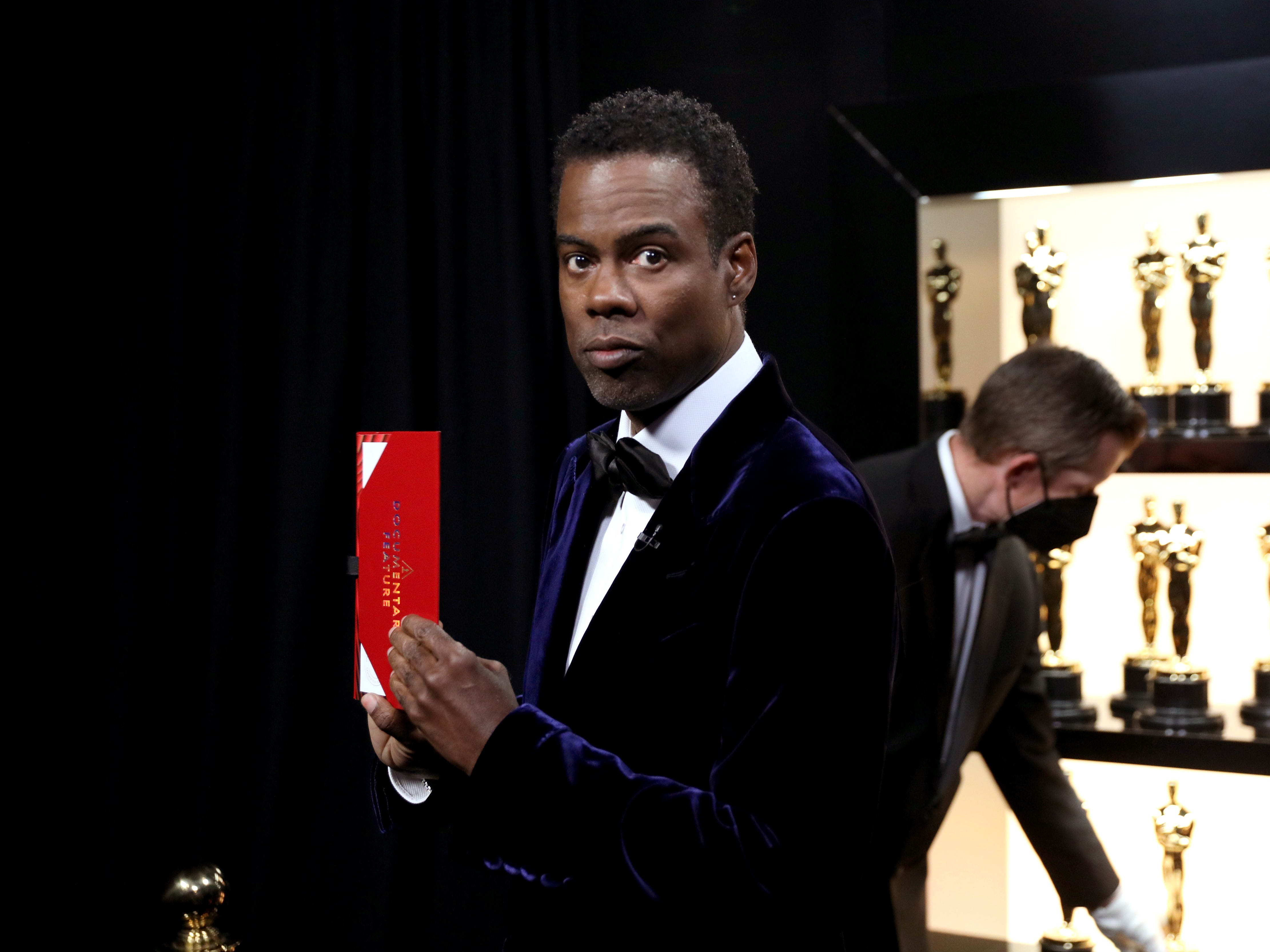 Chris Rock hält einen Oscar-Gewinner-Umschlag mit Oscar-Statuen hinter sich