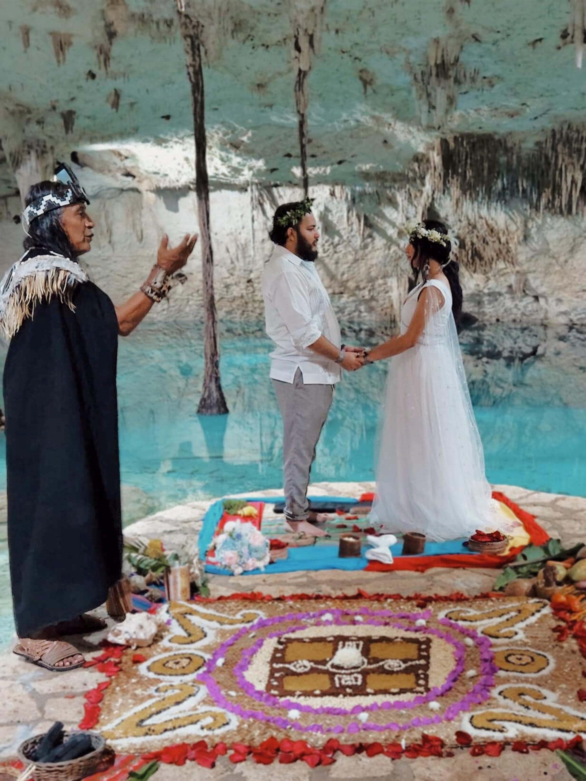 Eine Braut und ein Bräutigam halten Händchen vor einem Fluss, während ein Schamane ihre Hochzeit vollzieht.