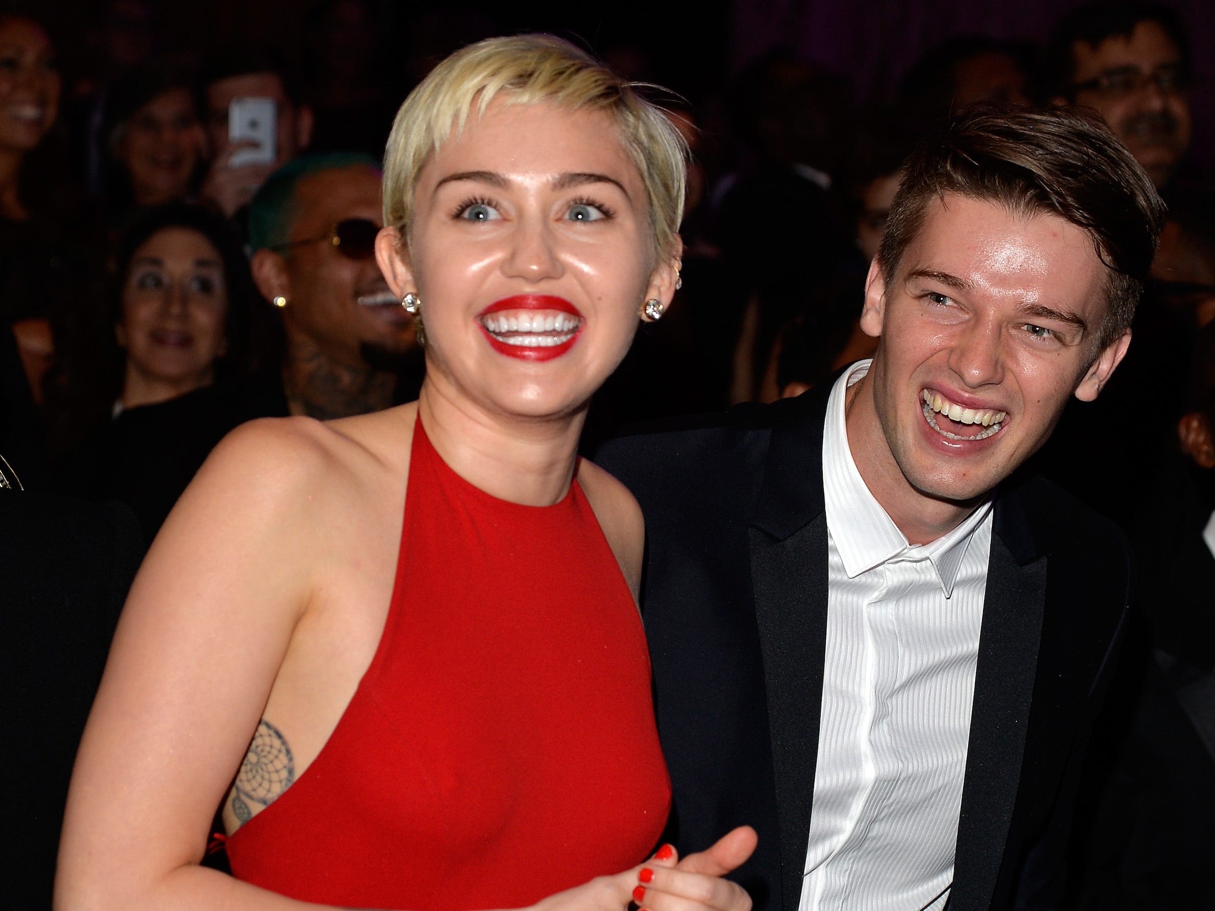 Miley Cyrus, in einem roten Kleid und Lippenstiften, lächelt während einer Veranstaltung mit Patrick Schwarzenegger, der einen schwarzen Anzug und ein weißes Hemd trägt.