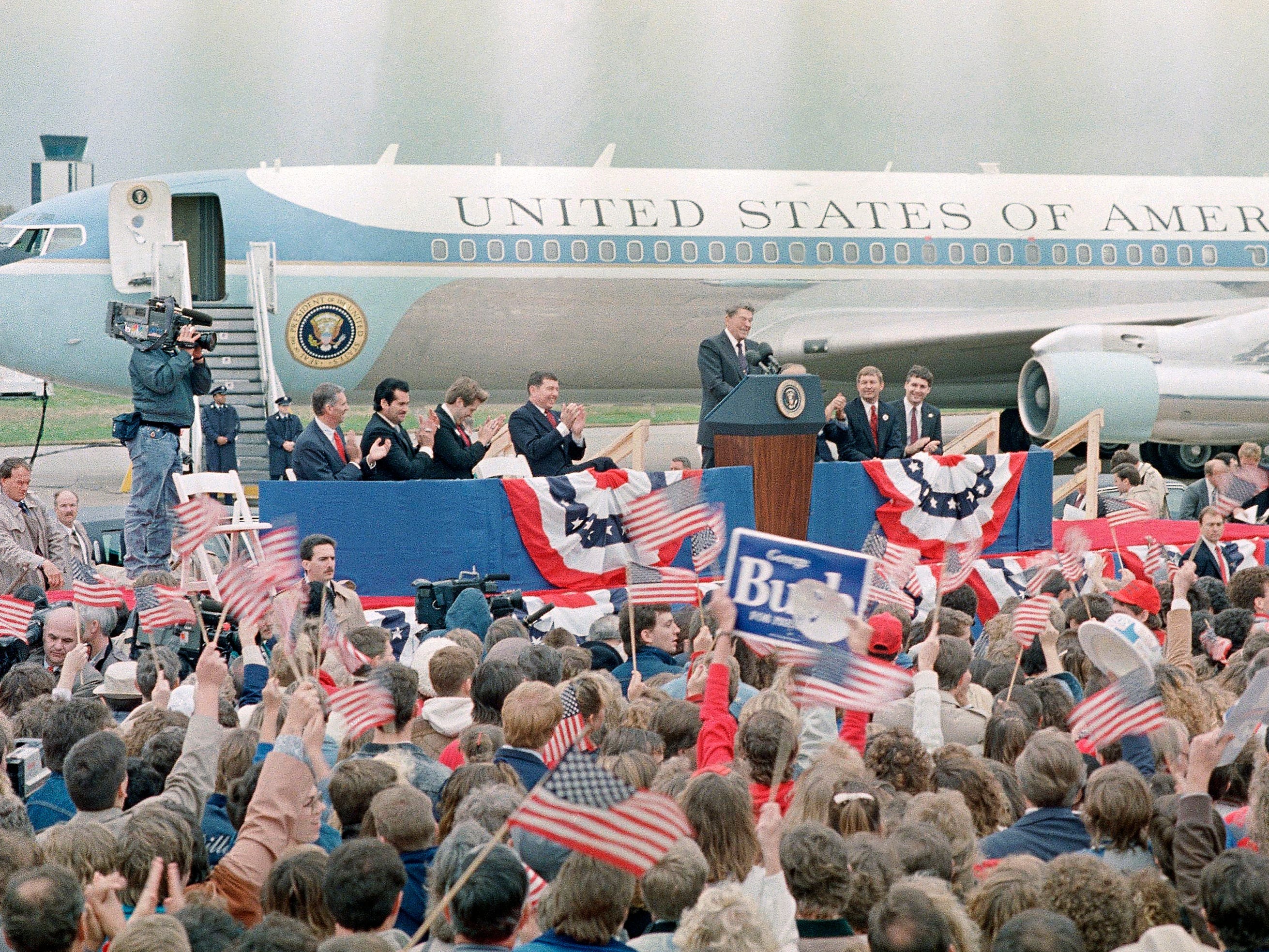 Eine Menschenmenge steht mit amerikanischen Flaggen und Gesichtern, dann Präsident Ronald Reagan, der hinter einem Podium vor einem blau-weißen Jet steht