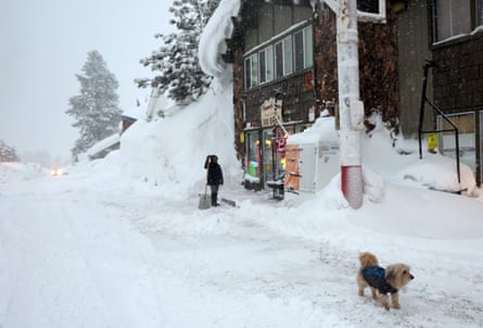 Eine Frau und ihr Hund stehen im Schnee vor einem Holzladen in einer Bergstadt.