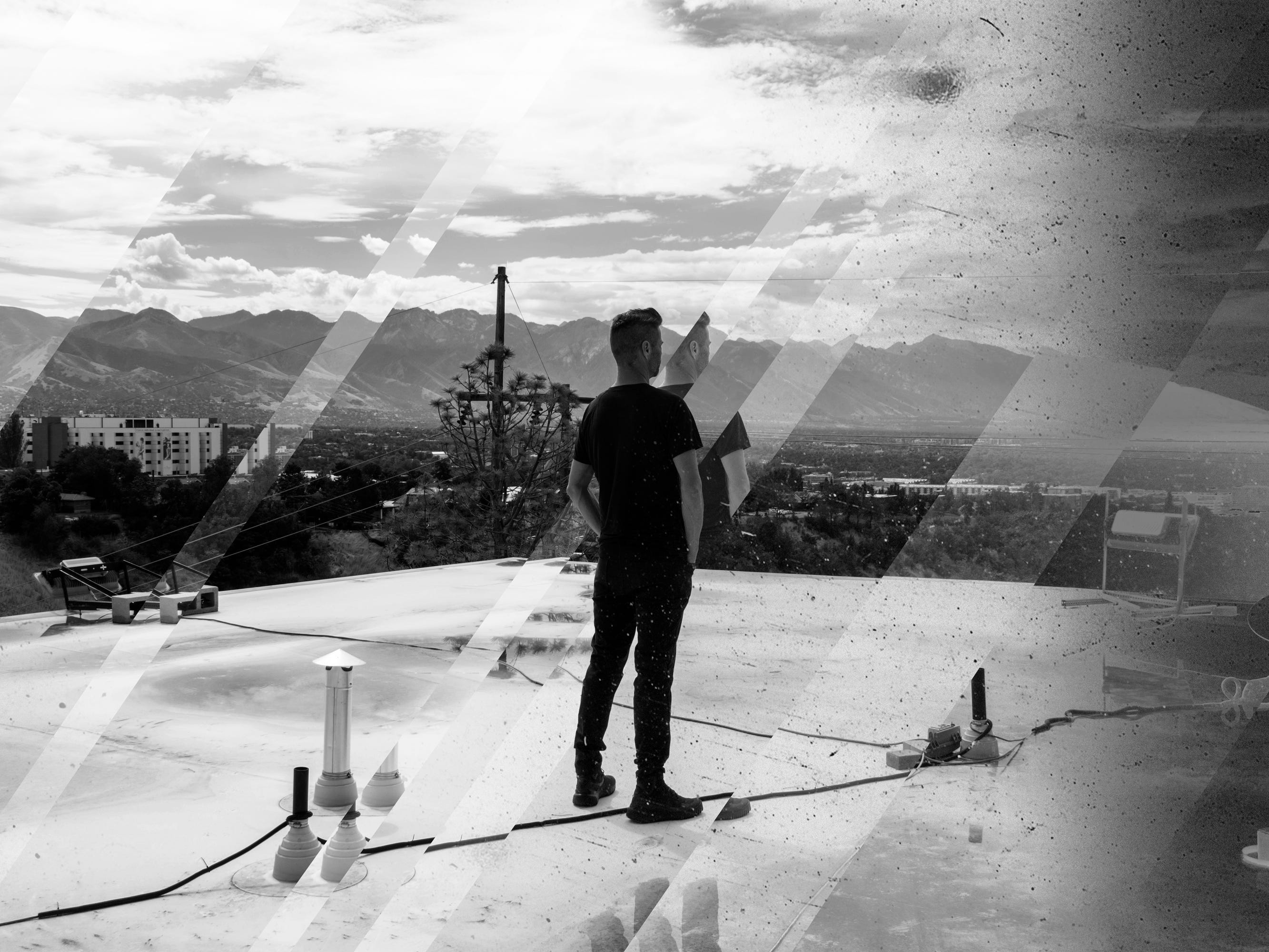 Schwarz-Weiß-Bild eines Mannes in einem schwarzen T-Shirt, der auf einem Dach steht und von der Kamera weg in Richtung einer Bergkulisse blickt.