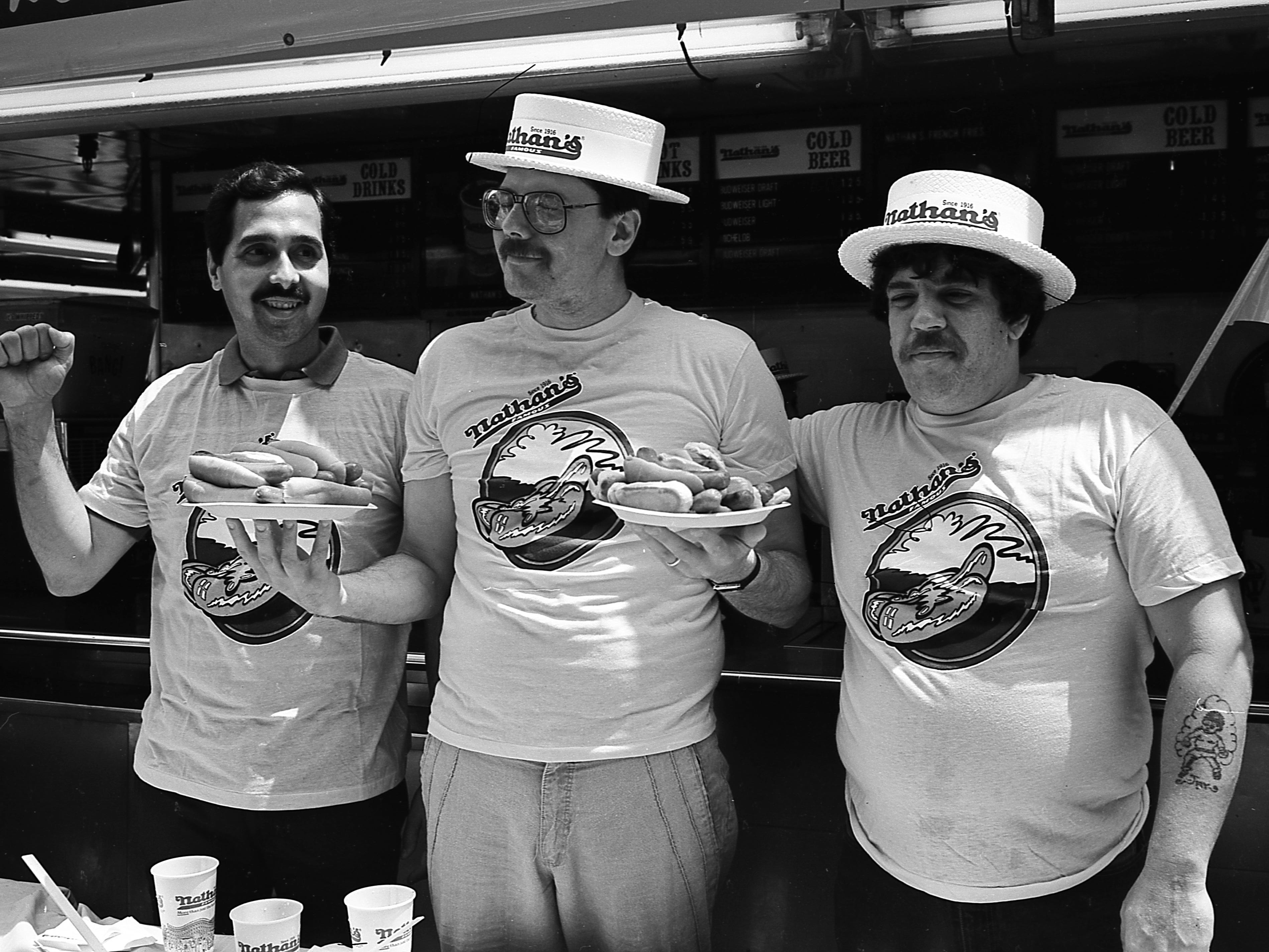 Porträt von drei nicht identifizierten Teilnehmern, die vor dem Nathan's Hot Dog Eating Contest 1987 auf Coney Island mit Hotdog-Tellern posieren