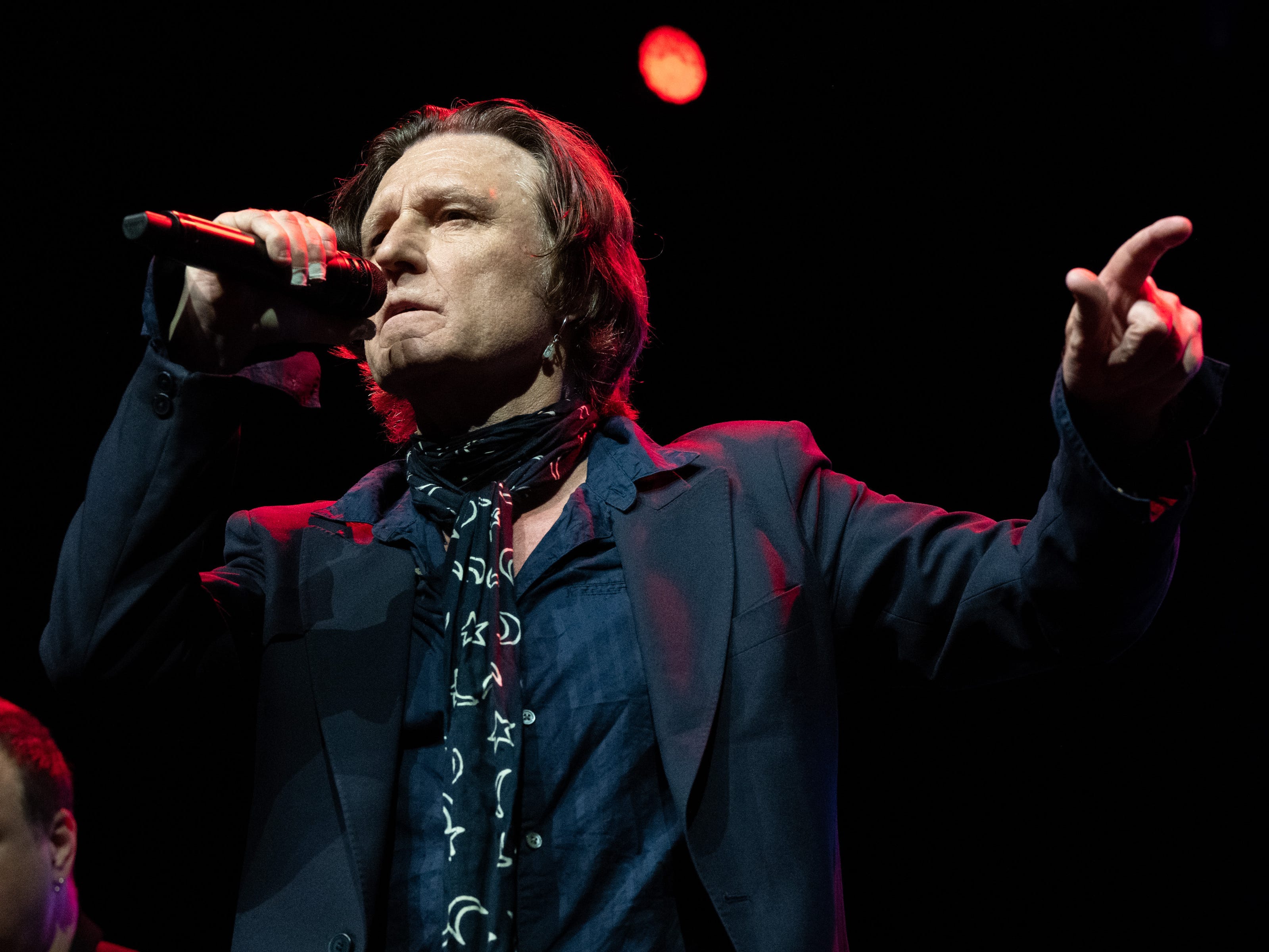 Sänger John Waite auf der Bühne, hält ein Mikrofon hoch und gestikuliert mit der linken Hand.  Er trägt ein dunkelblaues Hemd, einen schwarzen Blazer und einen schwarz-weiß gemusterten Schal