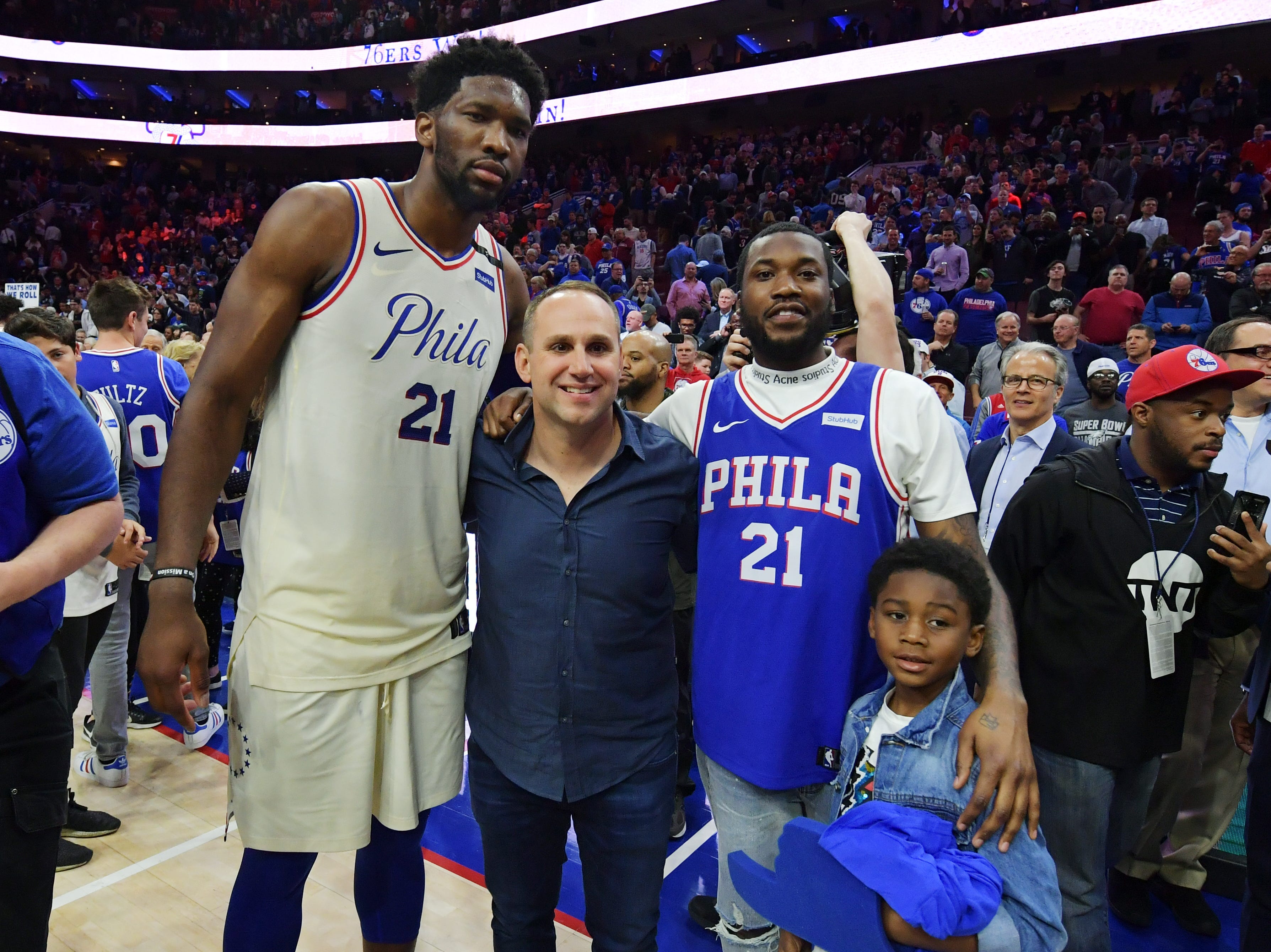 Joel Embiid, ein großer Mann im Philadelphia 76ers-Trikot, Michael Rubin, ein kleinerer Mann in einem blauen, weit ausgezogenen Hemd, Meek Mill, ein weiterer Mann im Philadelphia-Trikot, und sein Sohn Papi, ein kleines Kind, stehen auf einem Basketballplatz