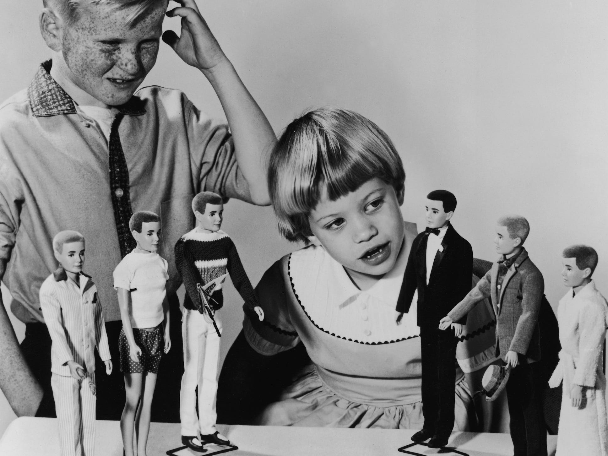 Ein kleiner Junge sieht verwirrt aus, während ein junges Mädchen 1961 Ken-Puppen inspiziert.