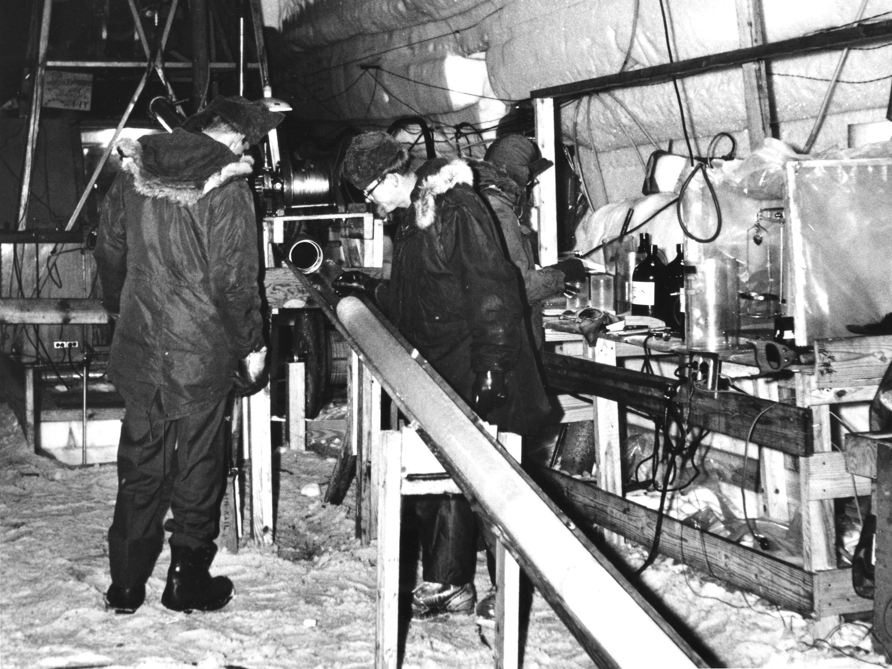 Ein altes Schwarz-Weiß-Foto von J.  Kasl und D. Garfield besichtigen einen geborgenen Kern im Camp Century im Jahr 1966. Die Forscher tragen dicke Daunenjacken mit Fellkapuzen und stehen neben komplex aussehenden Metalltischen mit wissenschaftlichen Maschinen.