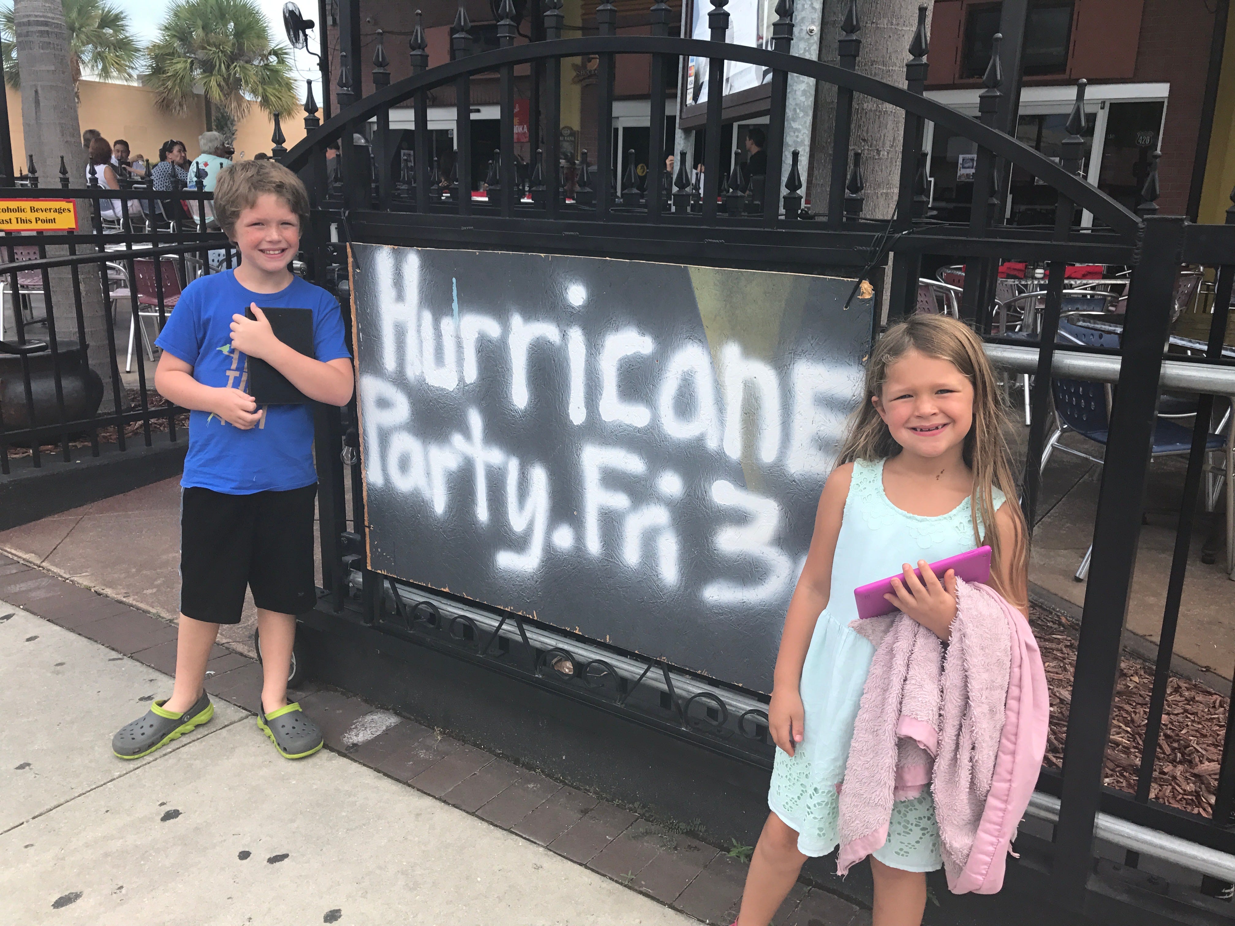 Hurrikan-Party am 3. Freitag, aufgesprüht auf einer Holzplatte auf einem Zaun, daneben stehen zwei Kinder
