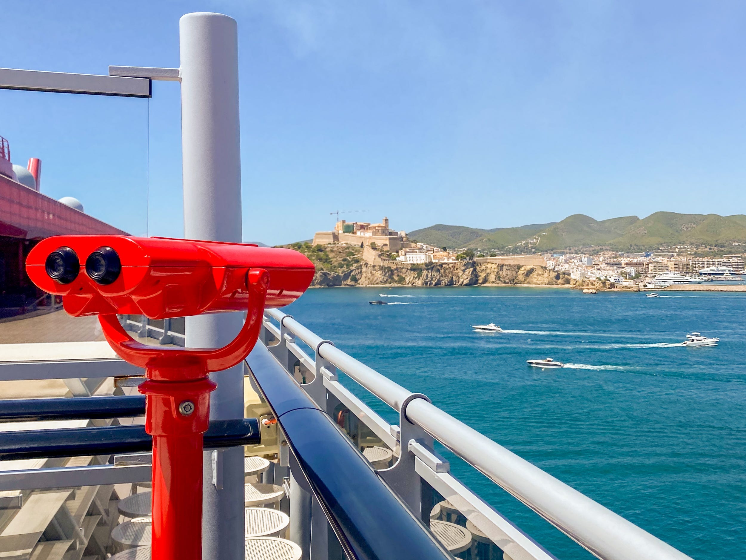 Links ein rotes Teleskop auf dem Oberdeck eines Kreuzfahrtschiffes mit dem Meer und einem fernen Hügel rechts.